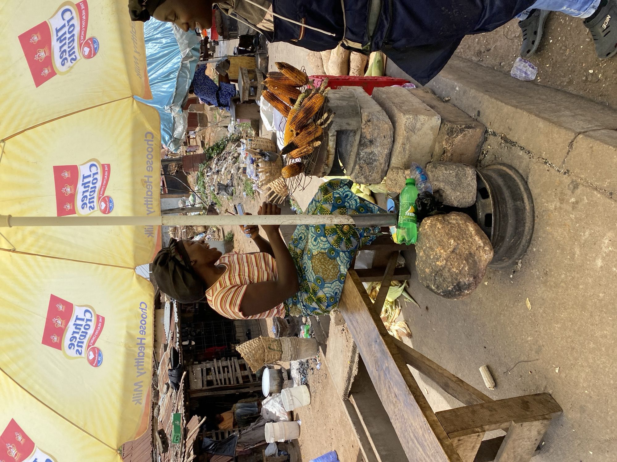 This media file captures a vibrant street vendor scene in Bukuru, Nigeria, oriented vertically from a slightly elevated perspective. The main subjects are two women and a market stall.
In the foreground, a woman is seated on a wooden stool, wearing a striped top and a patterned skirt. She appears to be the vendor, looking towards the right side of the frame, possibly observing passersby or another activity. Her hands are near her lap, holding what looks like a phone. Another person, partially visible at the top right, stands or leans near the stall, wearing a dark jacket and blue jeans, with "NICE" visible on their dark-colored footwear.
The central activity revolves around a street food stall, likely selling grilled corn. Several ears of roasted or grilled corn are displayed on a wire rack, positioned atop large stone blocks that form part of the stall's structure. A bright yellow umbrella, providing shade, dominates the left side of the image. The umbrella features reversed text, including "CLOMUD" (or similar brand name, possibly "KLIM" milk, often seen as "MILK" reversed), "THE HEALTHY CHOICE," and numerical text like "85" or "35," along with a small logo.
Notable objects include a green "TEEM" brand soft drink bottle, several white plastic buckets, woven baskets, and a discarded tire rim used as part of the setup. The setting appears to be a busy, outdoor market or roadside area, with concrete and dirt ground. In the background, there's an accumulation of discarded items, suggesting typical market refuse. The natural lighting indicates it is daytime, likely clear and sunny. The overall atmosphere suggests a casual, everyday commercial interaction.