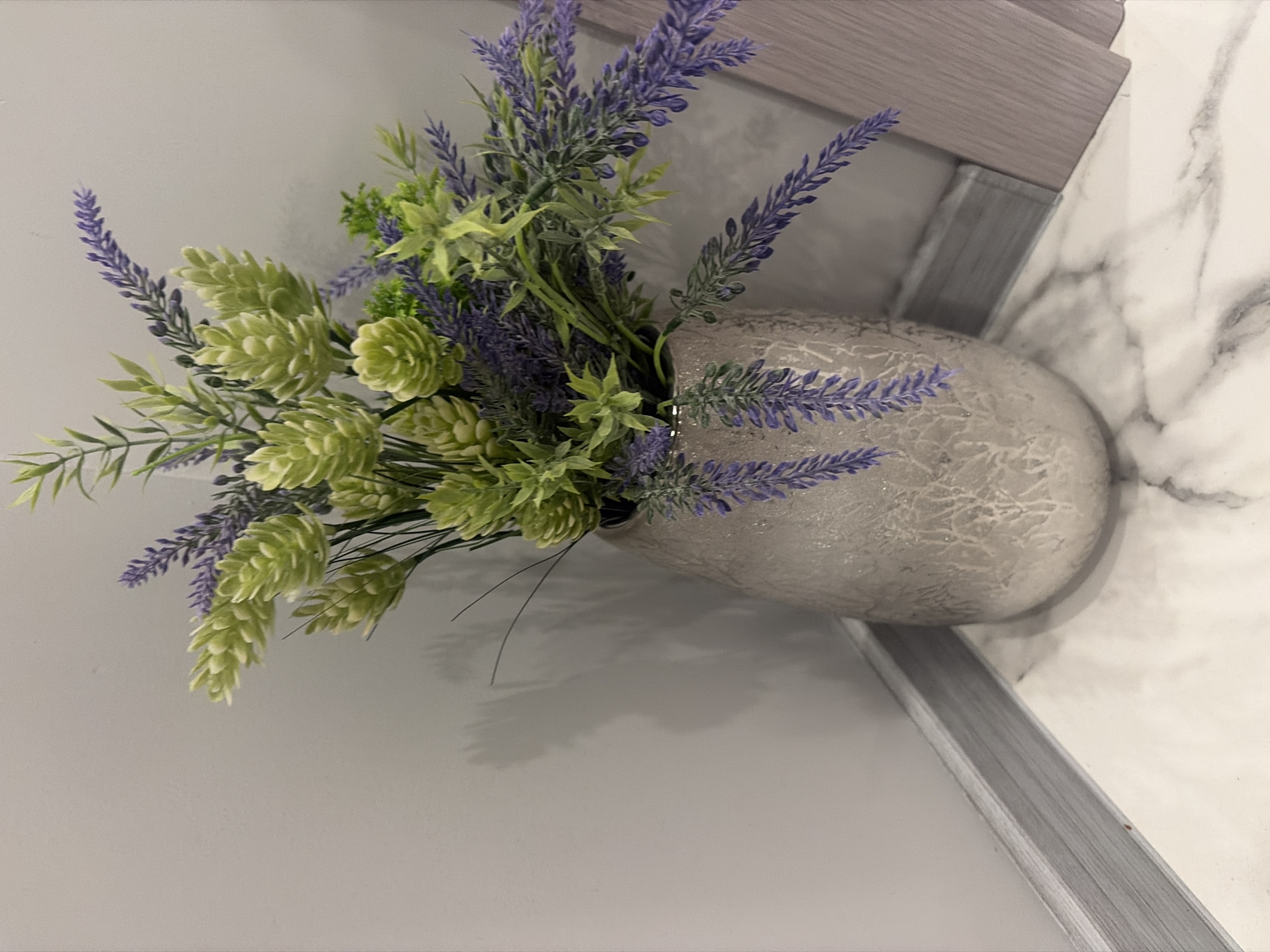 A still life photograph captures a decorative arrangement in the corner of a room. A light gray wall meets a light gray baseboard, which sits on a marble-patterned floor. In the corner, a textured, light beige vase holds a bouquet of artificial flowers. The bouquet features clusters of light green, cone-shaped blooms and sprigs of purple lavender. The lighting creates a soft shadow on the wall behind the vase. The scene is devoid of human presence or activity. The location is identified as Podgumer, Bulgaria.