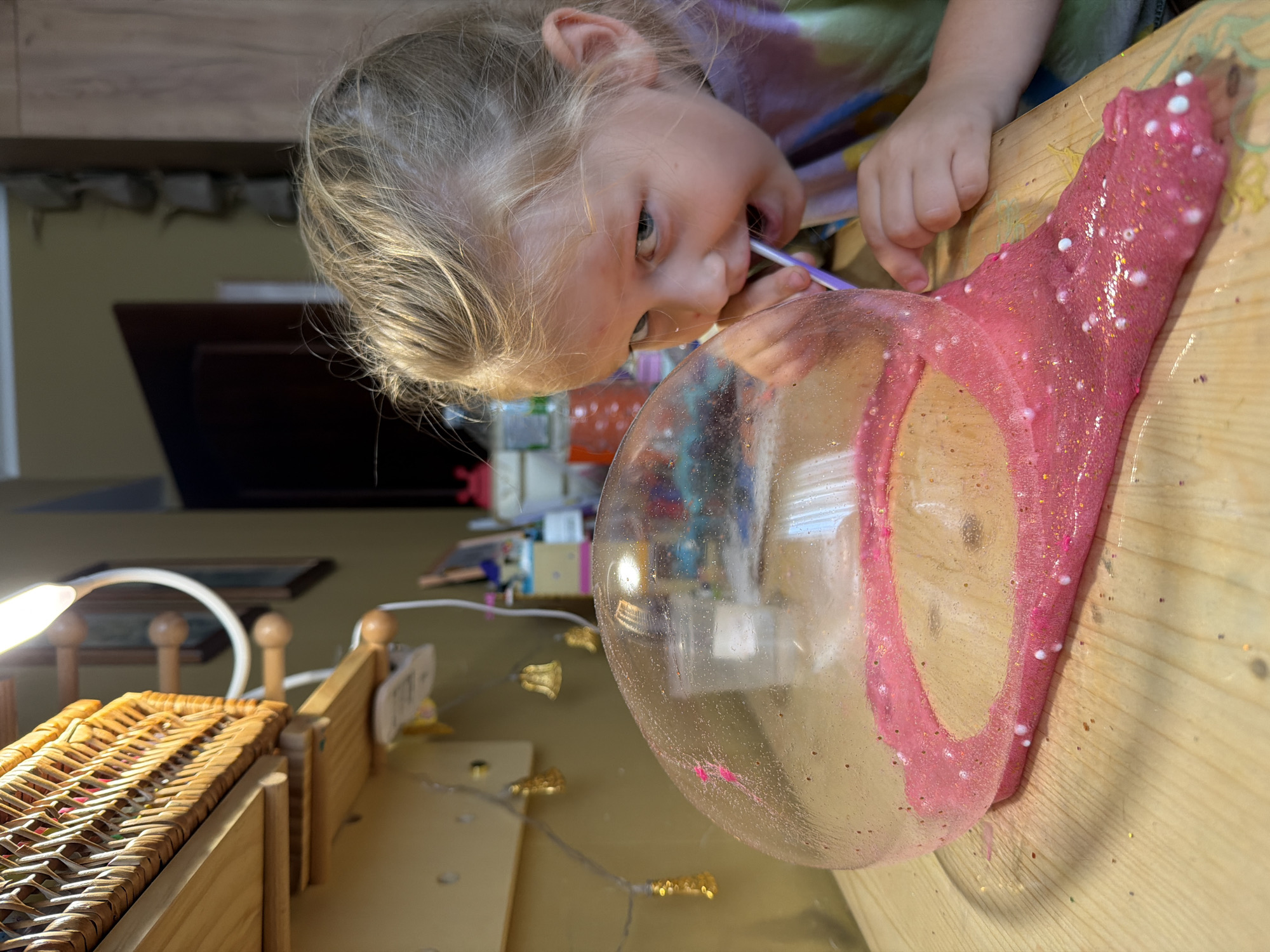 A close-up shot captures a young blonde girl with blue eyes, wearing a tie-dye shirt, leaning over a wooden desk. She is actively engaged in an activity with a large, clear bubble made of pink glitter slime. The girl is holding a purple straw to her mouth, seemingly blowing into the slime bubble. A portion of the bubble has detached and spread out on the desk, revealing its texture and glittery content. The desk also holds a few small white beads and scattered glitter. In the background, a wicker basket is visible on a wooden shelf, along with some decorative string lights featuring small golden bells. The overall environment suggests an indoor setting, possibly a home or classroom, with a neutral-colored wall and cabinetry in the background. The lighting suggests daytime. The image does not provide any visual cues to suggest a location in Khimki, Russia.