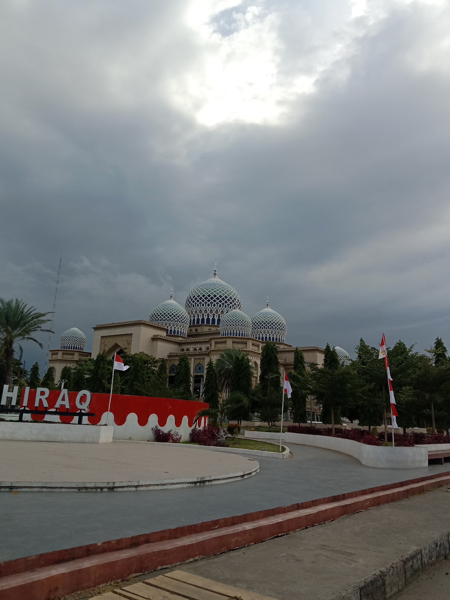 The image depicts a mosque with multiple domes in Lhokseumawe, Indonesia. The mosque features intricate blue and white patterned domes and has a light-colored facade with decorative carvings. In the foreground, a red and white sign with the text "HIRAQ" is visible, along with Indonesian flags planted on poles. Surrounding the mosque are lush green trees and palm trees. The sky is overcast with dark clouds, suggesting potential rain or a stormy weather condition. The scene is captured from an outdoor perspective, showcasing the architectural grandeur of the mosque and its surrounding landscape.