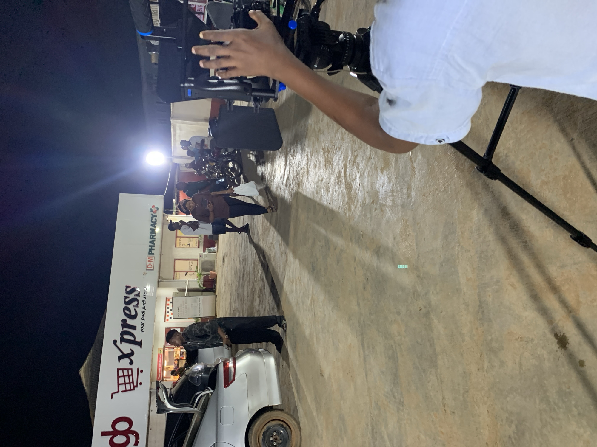 The image captures a scene at night in what appears to be Ikorodu, Nigeria. A "Go Xpress - Your Padi Padi Store" and "D&M Pharmacy" storefront is visible. Several people are present, including some near the storefront, and one individual loading items into the back of a silver car with the driver's side door open. The foreground is dominated by a camera operator filming the scene, with the camera mounted on a tripod. The ground is paved and shows signs of wear. A spotlight illuminates the scene. Other persons appear to be conversing. There are parked motorcycles nearby.