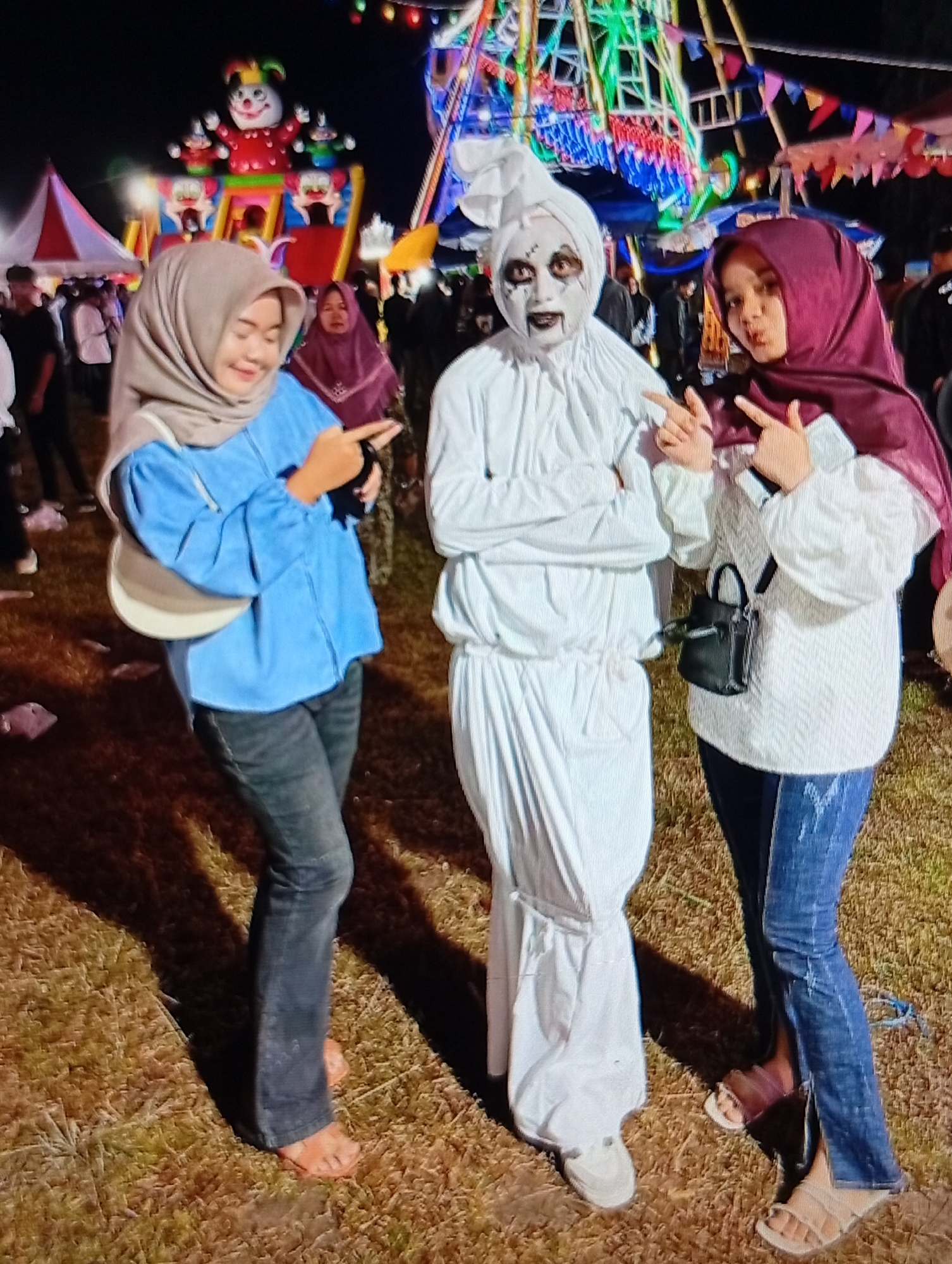 This image, taken in Lhokseumawe, Indonesia, depicts three individuals posing outdoors at night in what appears to be a carnival or night market setting.
In the foreground, from left to right:
A woman wearing a light beige hijab, a light blue long-sleeved shirt, and dark-colored jeans. She has a light-colored shoulder bag draped across her body. Her right hand is raised with the index finger pointing towards the right, and she is smiling while looking towards her right. Her right foot is bare on the ground, and her left foot is obscured.
The central figure is an individual in a white, shroud-like costume resembling a "pocong" (an Indonesian ghost wrapped in a burial shroud). The costume covers the entire body, including the head, which is cinched at the top. The face is painted white with heavy black detailing around the eyes and mouth, creating a ghoulish, skeletal appearance with dark lines and painted teeth. The individual's arms are crossed over the chest, and they are wearing light-colored athletic shoes.
To the right, a second woman is shown wearing a dark maroon hijab, a white textured long-sleeved sweater, and dark blue jeans with vertical slits at the ankles. She has a small black shoulder bag worn across her body. Both of her hands are raised, making 'peace' signs (V-gestures) with her index and middle fingers. She has a slight pout and is looking directly at the camera. She is wearing light-colored sandals.
The background features a lively night scene with illuminated amusement park attractions. Visible elements include a large, colorful inflatable bouncy castle or play structure adorned with cartoon figures (including a clown character) in the upper left. To the right of this, a brightly lit Ferris wheel or similar ride with multicolored lights (green, blue, red) is prominent. Various strings of colorful flags or pennants are strung across the upper portion of the background. Blurred figures of other people and structures are visible in the distance. The ground beneath the individuals is dry, light brown, disturbed earth or sparse grass. The overall lighting suggests an evening or nighttime setting.