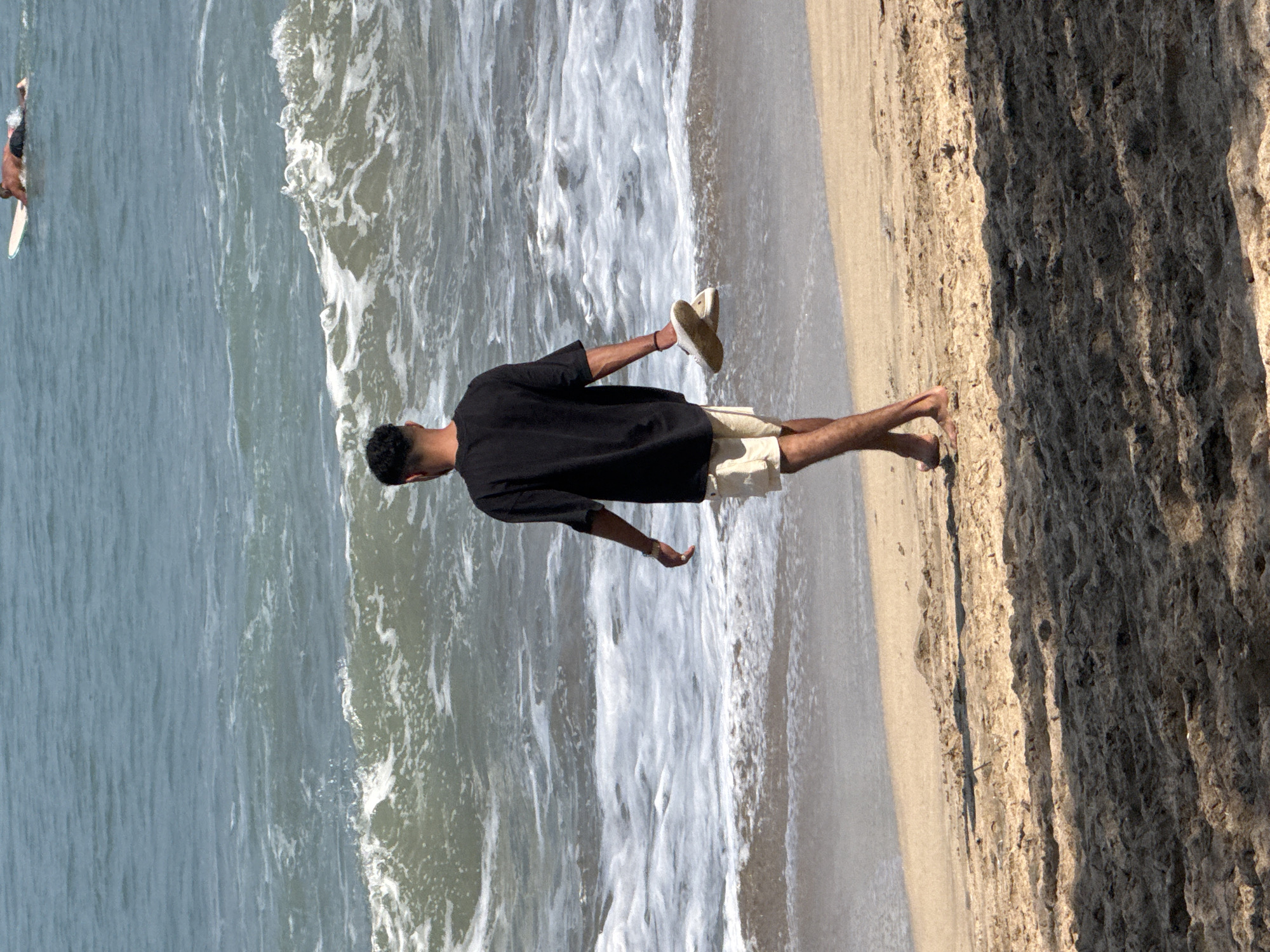 In Kuta, Indonesia, a young man with dark, curly hair walks on a sandy beach, holding a pair of white sandals in his right hand. He is wearing a black t-shirt and light-colored shorts. The ocean waves are breaking behind him, with white foam spreading across the water's surface. In the distance, a surfer is lying on a surfboard in the blue ocean. The foreground shows the texture of the sand with dappled shadows, suggesting a sunny day.