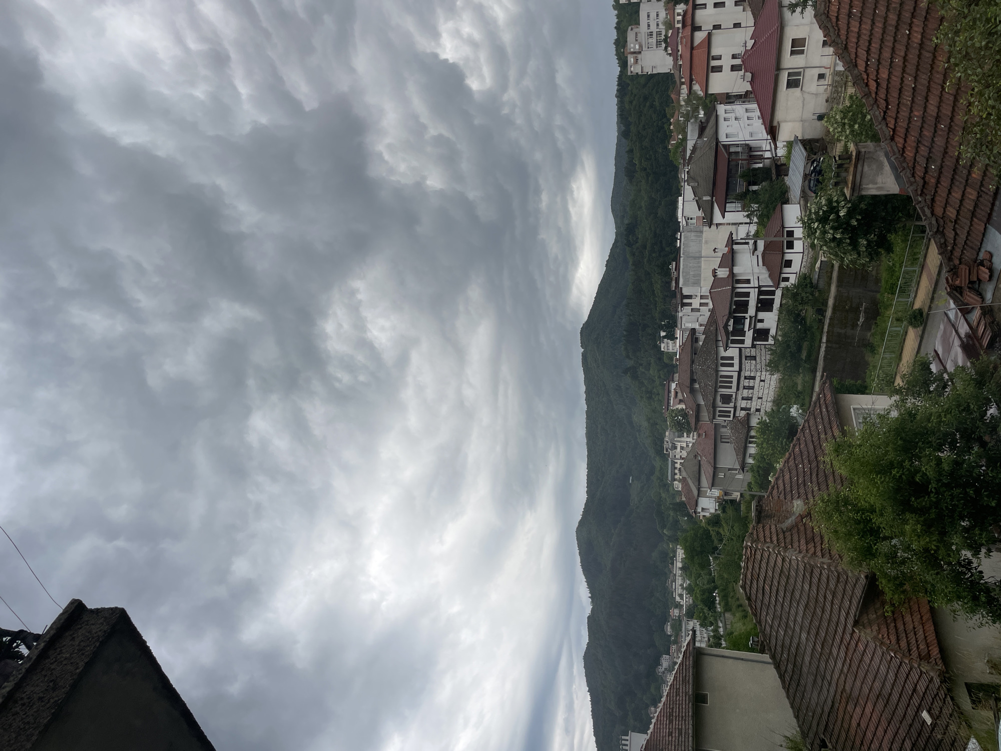 The image depicts a view of Zlatograd, Bulgaria, under a cloudy sky. In the foreground, the red-tiled roofs of several houses are visible, along with some greenery. Further into the scene, a cluster of buildings with white facades and dark roofs can be seen, some featuring traditional architectural elements. Beyond the town, a dense, tree-covered mountain range dominates the background. The sky is filled with heavy, grey clouds, suggesting an overcast or potentially stormy day. There are no people or specific actions captured in the image.