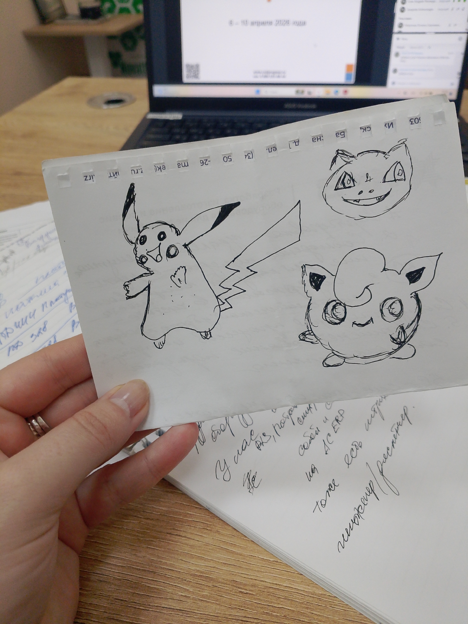 A hand holds a paper with drawings of Pikachu, Meowth, and Jigglypuff. The paper is placed on top of a notebook with handwritten text in Russian. A laptop is visible in the background. Text in the background reads "4-10 April 2026". The top of the page in the photo has text in a small font that references a city named Izhevsk, Russia.