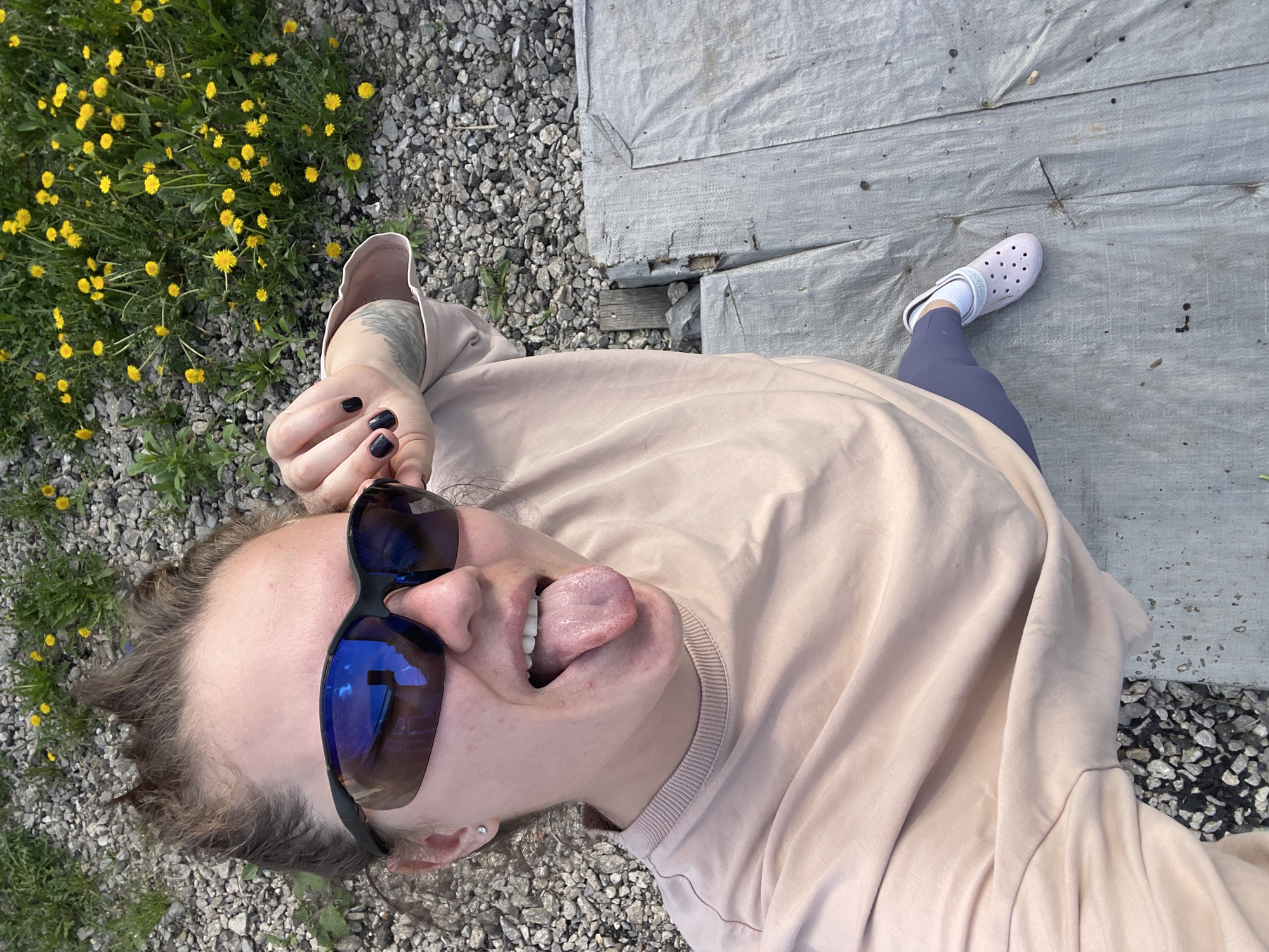 The image shows a person lying on the ground, seemingly on a path with gravel and dandelions on one side and a covered surface (possibly a tarp) on the other. The person is wearing sunglasses and sticking out their tongue, posing for the camera. They have on a light pink long-sleeved shirt, blue leggings, and a white clog-style shoe on one foot. The location is Misaylovo, Russia, based on the information provided.