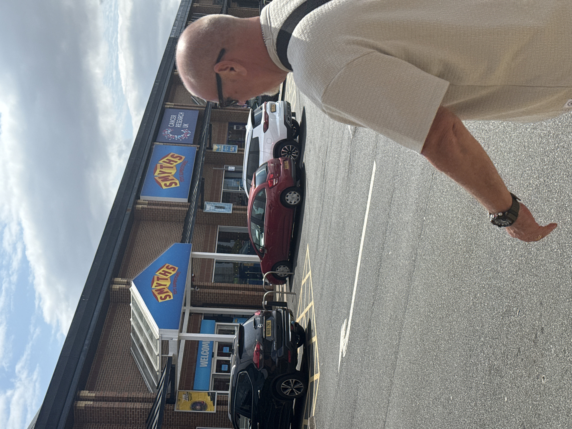 The image depicts the exterior of a Smyth's Toys Superstores location in Darlington, United Kingdom. In the foreground, a bald man wearing sunglasses and a light-colored textured shirt is partially visible, with his right arm and shoulder in the frame. He is wearing a watch on his left wrist. The background shows the brick facade of the Smyth's store, with prominent blue signage featuring the Smyth's logo. Adjacent to the Smyth's sign, there is a sign for "Cancer Research UK". The entrance to the store has a blue awning with the word "WELCOME" written in white. Several cars are parked in the lot, including a black SUV with the license plate "NU69 ZKE" and a red hatchback. The ground is asphalt, marked with yellow parking lines and a white arrow. The sky is partly cloudy.