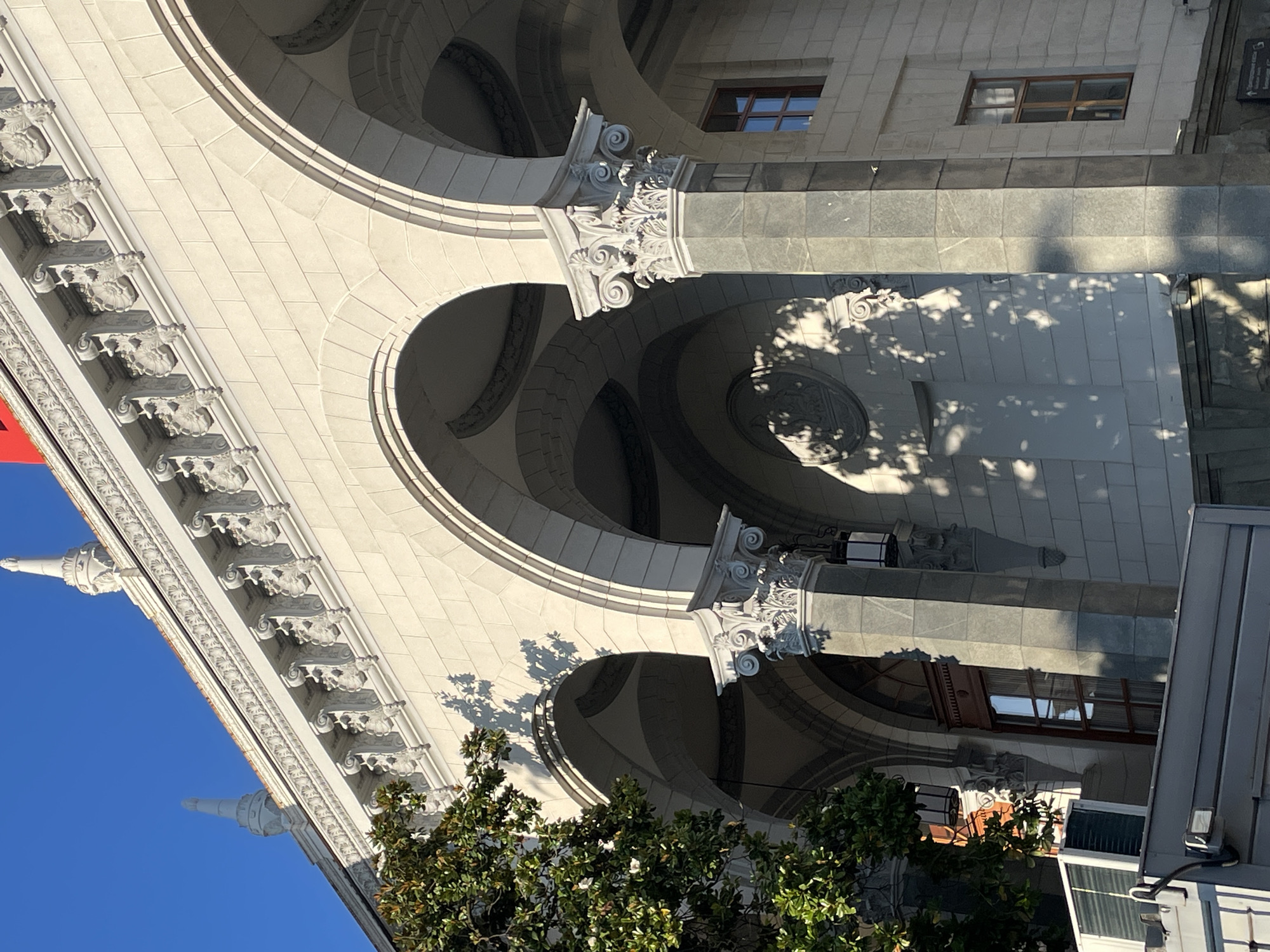 The image shows the exterior of a light-colored, ornate building with arched walkways supported by decorative columns. The architecture features intricate stone carvings along the roofline and on the column capitals. A portion of a large green tree is visible on the left side of the frame, casting dappled shadows on the building's facade. The sky above is clear and blue, indicating a sunny day. In the foreground, a modern air conditioning unit is mounted on a grey structure, and part of a window with a wooden frame is visible on the building. The building appears to be in an urban setting, likely in Sochi, Russia, given the architectural style which is reminiscent of Soviet-era public buildings.