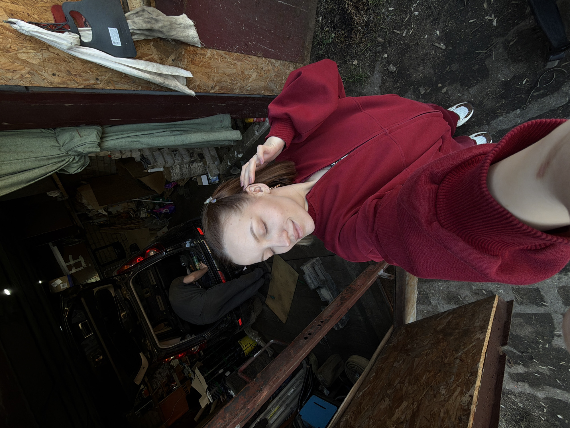 A person wearing a red zip-up sweatshirt stands next to an open garage or workshop in Reshetnikovo, Russia. A second person is visible inside the dark interior of a black van. The garage contains various stored items and tools. A section of the garage's exterior wall is constructed of plywood.