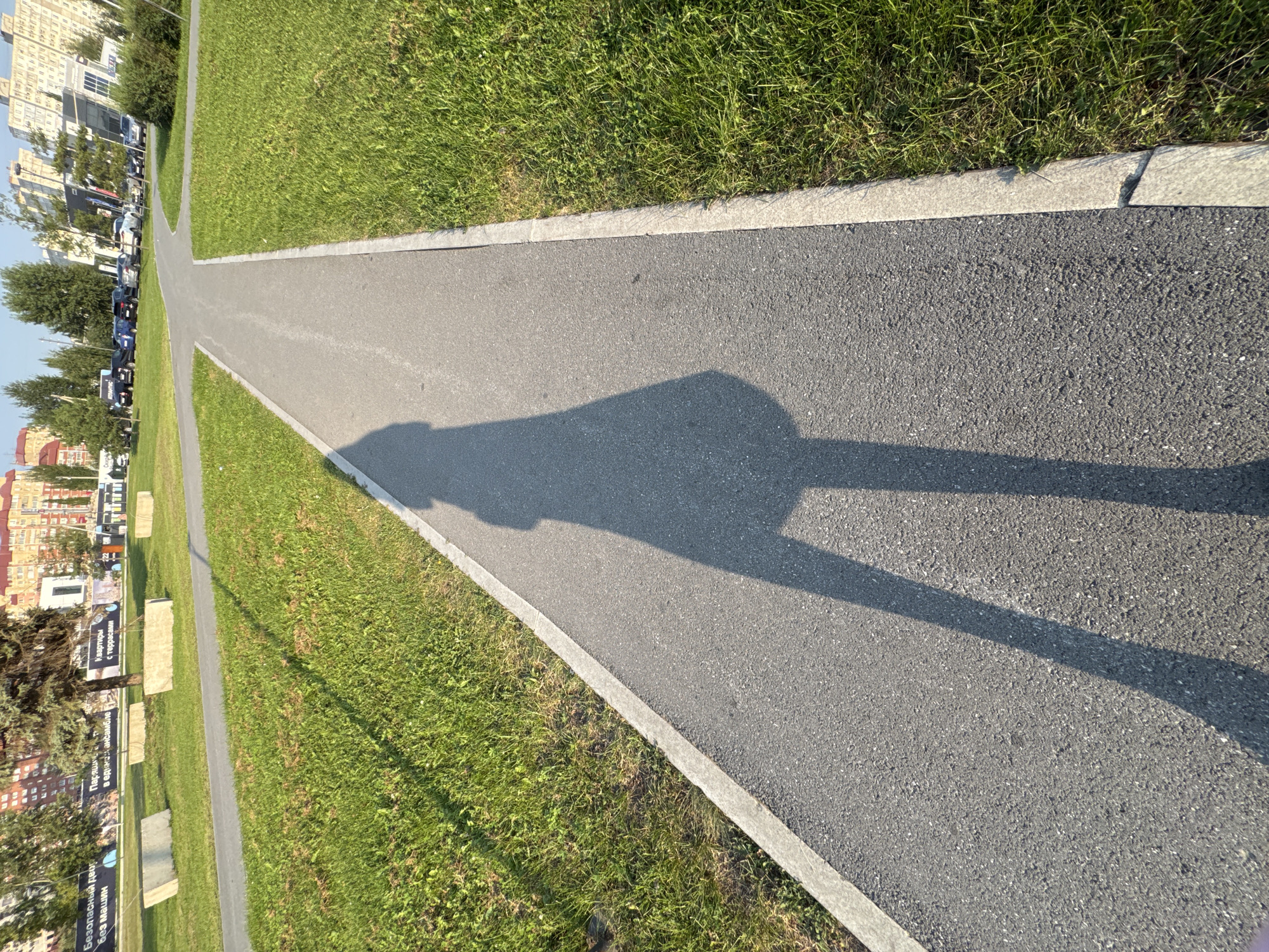 A long shadow of a person is cast across a paved pathway in a city park in Ufa, Russia. The pathway curves gently, bordered by a strip of concrete and then a well-maintained green lawn on either side. In the background, apartment buildings and trees are visible under a clear sky. Several concrete barriers are placed on the grass. The scene appears to be captured during daylight hours, likely late afternoon due to the elongated shadows.