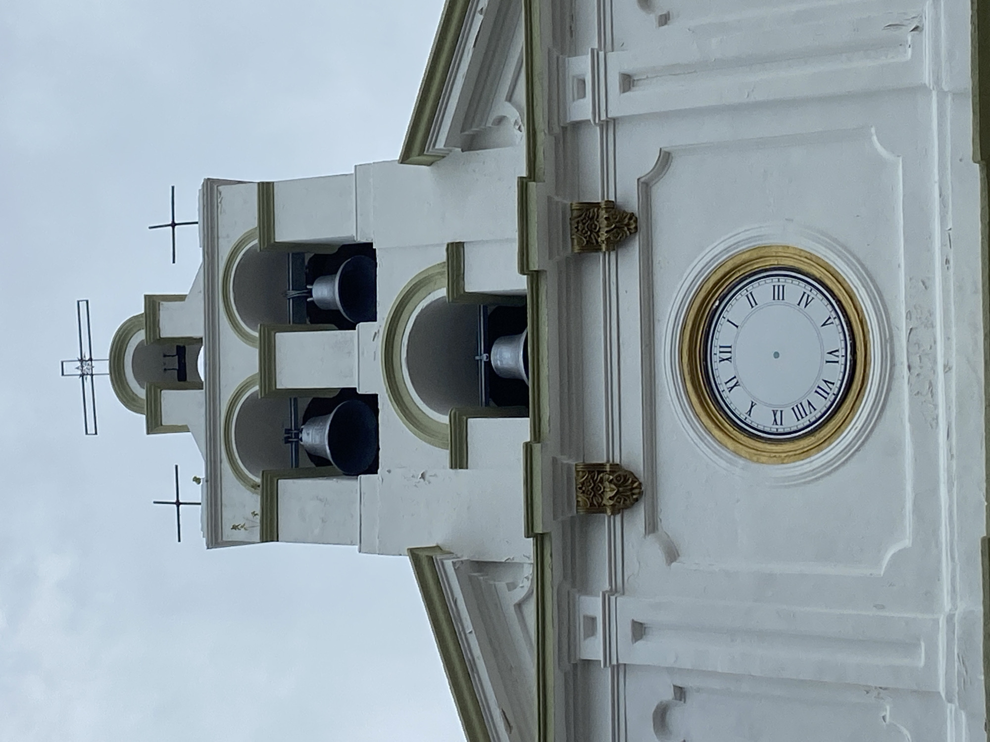 The image shows a close-up of a white church tower, possibly located in Río Chico, Venezuela, featuring a clock and bells. The tower has a three-tiered structure. Each tier features arches containing a bell. The clock face is circular with gold trim and Roman numerals. Two ornate, gold-colored embellishments are positioned on either side of the clock. A cross is visible at the top of the tower. The sky is overcast.
