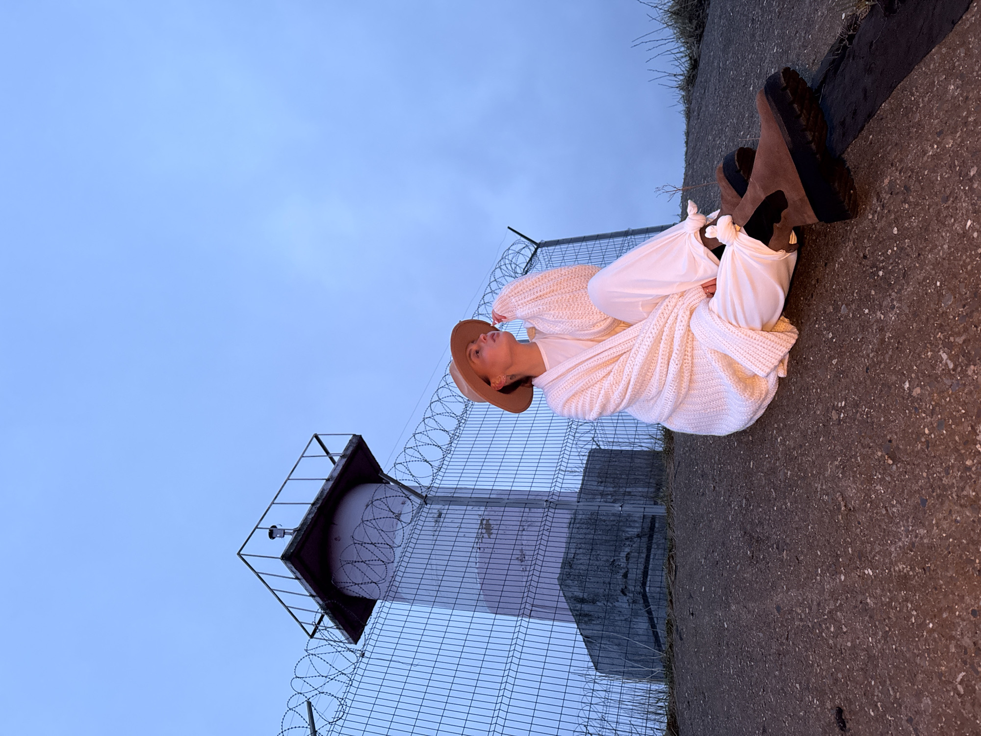 The image, oriented incorrectly, shows a person seated outdoors at what appears to be dusk or twilight. The sky is a muted, light blue-grey, suggesting an overcast evening.
A lone individual is the central subject, seen from a slightly low angle. They are wearing a light-colored, chunky knit cardigan or sweater, light-colored trousers that are gathered or tied at the ankles, and dark brown platform or wedge boots. A light brown fedora-style hat is worn on their head. The person is seated on dark, textured ground, possibly asphalt or concrete, leaning back slightly and looking upwards with a serene or contemplative expression.
In the background, a tall, slender structure resembling a tower or mast is visible, partially obscured by a chain-link fence. The fence is topped with multiple rolls of razor wire, indicating a potentially restricted or industrial area. The tower has a small, square platform at its apex, with what appears to be a light or surveillance camera fixture attached. The scene depicts an outdoor setting, possibly an industrial or perimeter area in Chaikovsky, Russia. There is no visible text in the image.