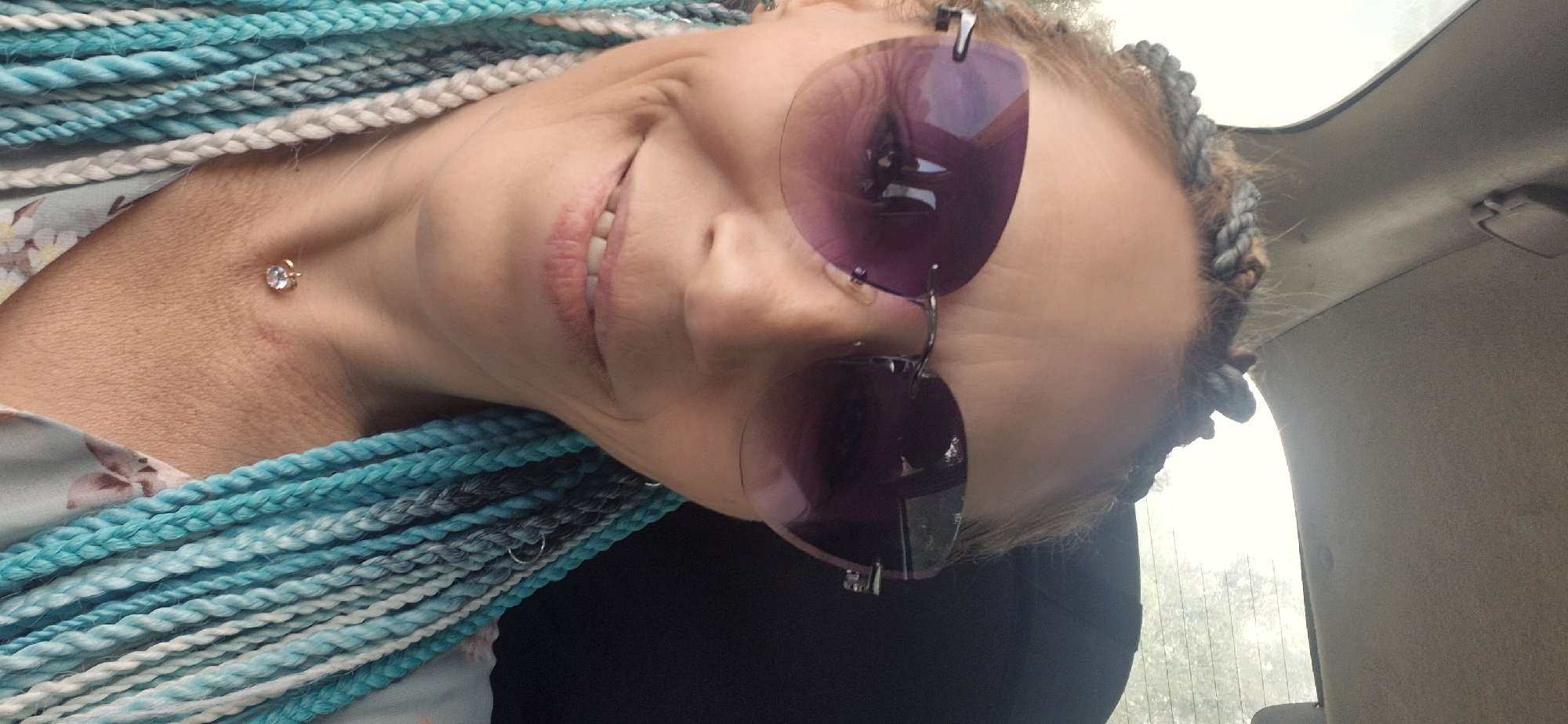 The media file is a close-up, tilted selfie of a woman with numerous braided extensions in her hair, featuring shades of blue, white, and gray. She is wearing large, round, purple-tinted sunglasses. The woman is smiling, with her lips slightly parted, revealing her teeth. A small, clear gemstone is visible as a piercing on her neck, just below her jawline. She is wearing a light-colored top with a floral pattern. The background suggests the interior of a vehicle, with a gray car headliner visible in the upper right portion of the frame. The overall lighting and the bright sky seen through a window behind her indicate it is daytime, likely outdoors, though the specific weather conditions are not discernible. The image focuses on the woman's face and upper body, with the braids prominently framing her.
