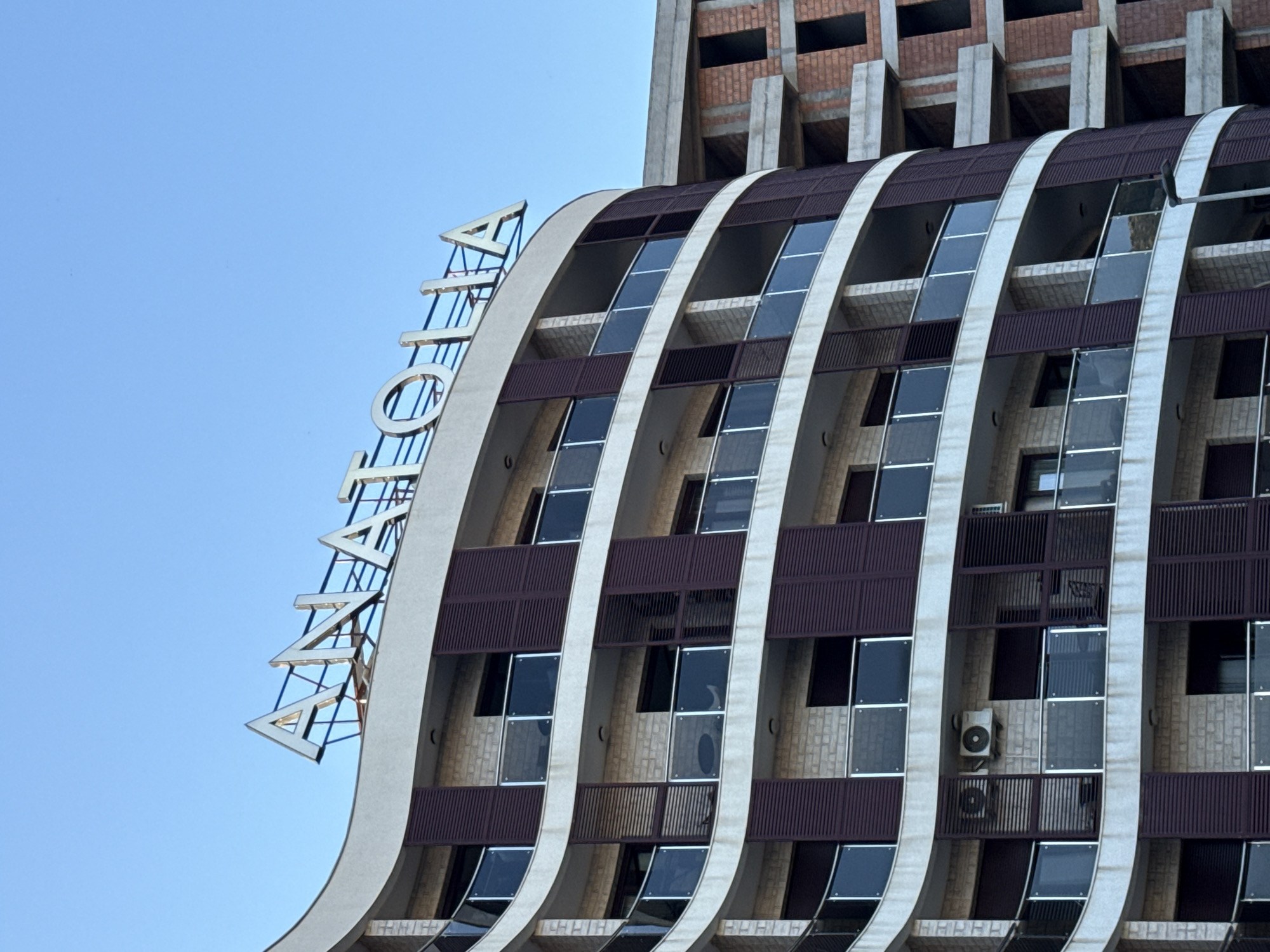 The image shows the exterior of a modern apartment building with a curved facade in Boreti, Montenegro. The building has multiple floors with balconies featuring glass railings. Dark colored shutters are installed on some windows. A large metal sign spelling out "ANATOLIA" is mounted on the rooftop. To the right of the main building, a partially constructed building is visible, made of brick and concrete. The sky is clear and blue. There are no people or animals visible in the image.