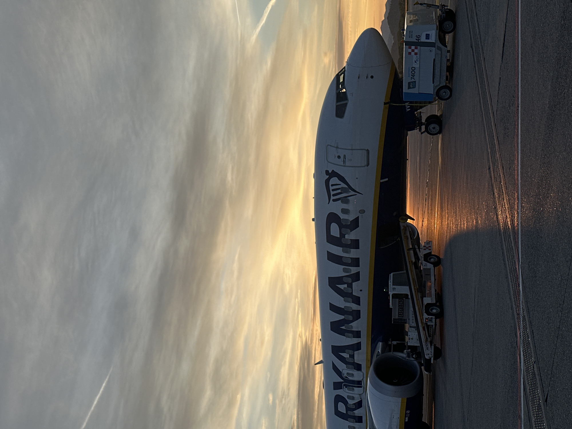 A Ryanair airplane is parked on the tarmac at Orio al Serio Airport, Italy. The plane is being serviced by a baggage truck and an Easy Load truck, with an orange sunset sky in the background.