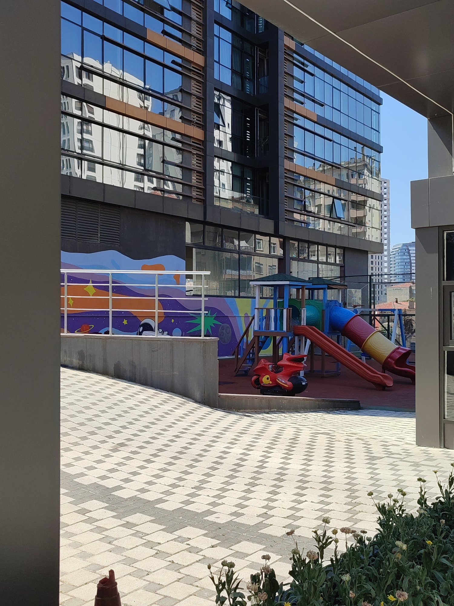 This is an outdoor, eye-level shot of a children's playground adjacent to a modern building in Kadıköy, Turkey. The building, with its dark facade and large, reflective glass windows, dominates the background. Reflections of other buildings are visible in the glass. In the middle ground, a colorful playground structure features slides, a climbing frame, and a small red toy vehicle. A mural depicting outer space adorns the wall behind the playground. The foreground consists of a patterned paved walkway and a planter with wilting flowers. The scene is bathed in daylight.
