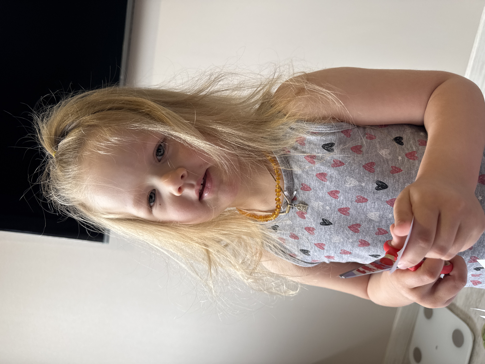 A young blonde-haired child is captured in an indoor setting, looking towards the viewer with a soft smile. The child has fair skin and light-colored eyes. Her long, light blonde hair is somewhat disheveled, with a yellow hair tie visible at the back of her head, gathering some strands. She is wearing a sleeveless grey top patterned with numerous hearts in pink, black, and white outlines. Around her neck, she wears a delicate amber beaded necklace from which a small silver-colored charm hangs.
The child is actively engaged, holding red-handled scissors in her right hand and a small piece of white paper in her left, appearing to be in the process of cutting. Her gaze is directed towards the camera or slightly beyond it. The background features a light-colored wall, a dark rectangular object (possibly a television or furniture) to the left, and a white, flat appliance with circular elements, resembling a weighing scale, on the floor to the right. The indoor lighting suggests a comfortable home environment. The child's expression conveys a sense of engagement and calm. No visible text is present in the image.