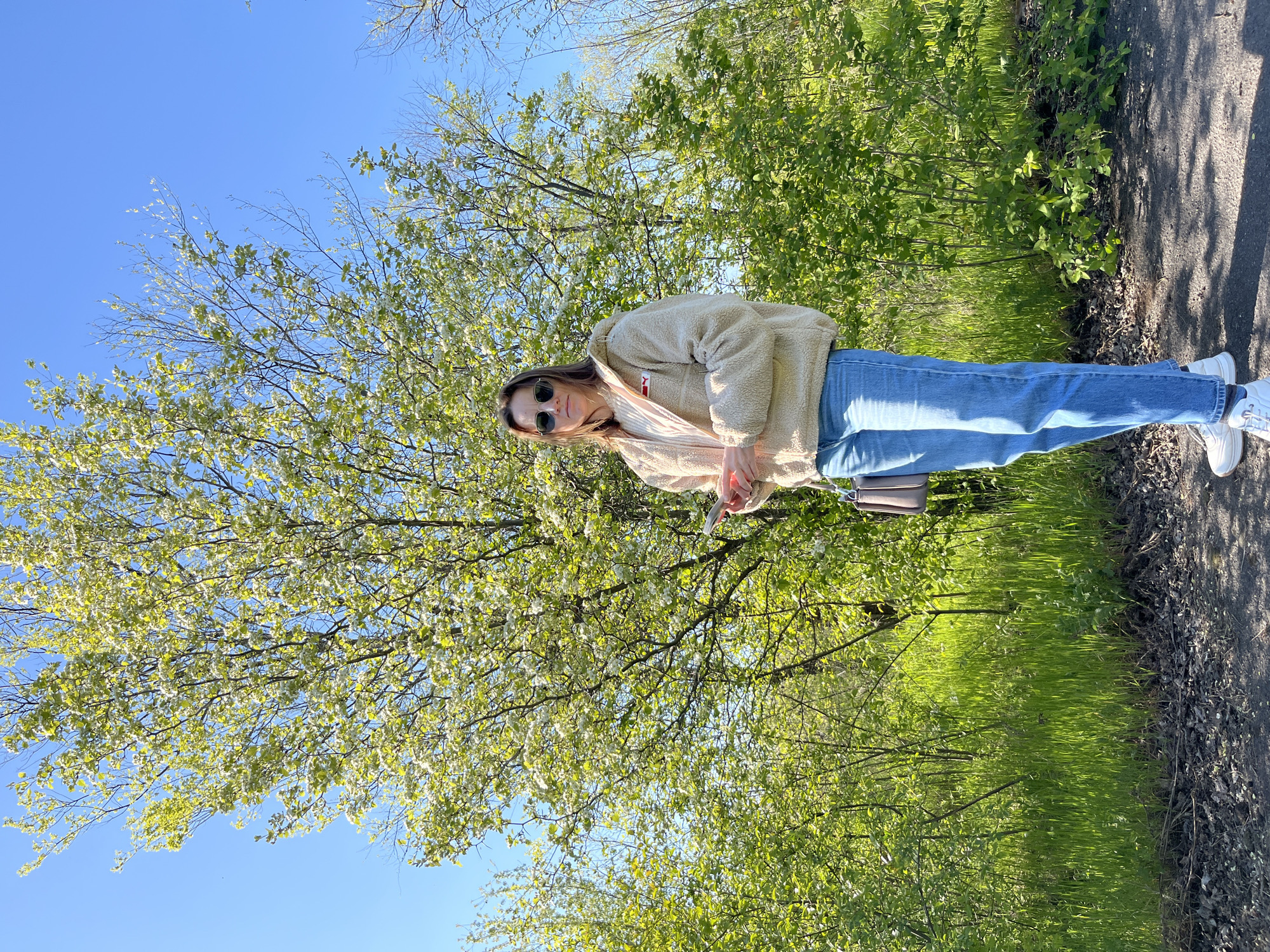 A young woman stands on a path next to lush green vegetation and a tree in full bloom, set against a clear blue sky. The location is likely a park or natural area, based on the dense greenery and blooming tree. The woman is wearing a beige jacket, blue jeans, and white sneakers, and she has sunglasses on. She holds an object, potentially a phone, in her hands. The path, covered with leaves and debris, leads to a paved area. The environment suggests a sunny day in the spring or early summer season. The image was taken at a slight angle, tilted counter-clockwise.