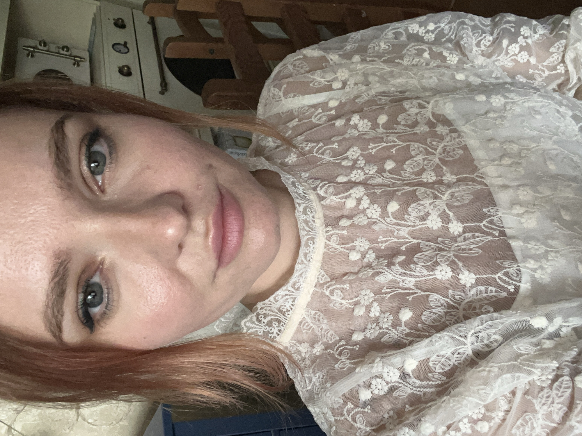 The media file is a close-up, low-angle portrait of a young Caucasian woman with light skin and reddish-brown hair. She is wearing a sheer white lace top with floral embroidery. Her left eye is visible, showing a light blue iris and dark eyelashes. Her lips are slightly parted, and she has a neutral expression. The background is out of focus, but appears to be an indoor setting with wooden furniture and what looks like a kitchen appliance in the upper left corner. There are no visible text elements in the image. The lighting suggests it is daytime.