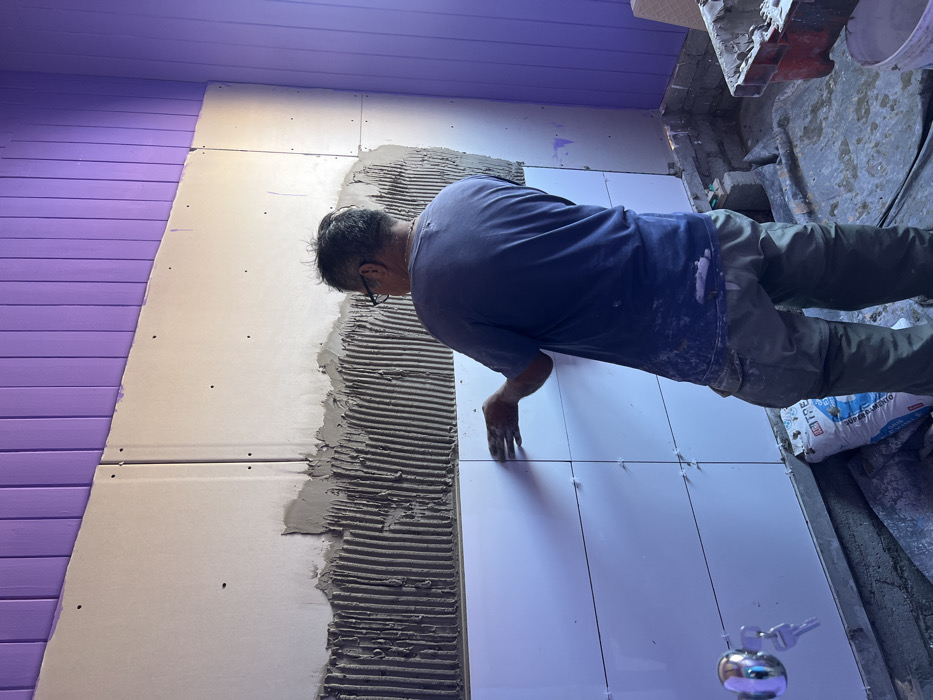 The image captures a scene of a person actively involved in tiling a wall, likely in an indoor construction or renovation setting. The perspective is from slightly above and behind the person, angled to the right.
The central figure is an adult, appearing to be a male, seen from his back and right side. He is wearing a dark blue short-sleeved t-shirt and olive green or khaki-colored long pants. His clothing shows smudges or splashes of white material, likely from the tile adhesive or grout. He has short, dark hair and is wearing glasses. He is leaning forward, focused intently on the wall he is working on, with his right hand positioned near a newly placed tile.
The wall itself is composed of different sections. To the left, there is a section of horizontally-oriented wooden planks or siding painted a vibrant lavender or light purple color. This section meets a lighter-colored substrate, possibly drywall or cement board, which forms the surface for the tiling. This substrate has several small screw or nail holes visible.
On the lighter wall section, a thick layer of gray tile adhesive has been applied in textured horizontal ridges, indicating it was spread with a notched trowel. To the right of this prepared section, several shiny white, rectangular tiles have already been installed. Small, white plastic tile spacers are visible between the tiles, ensuring uniform gaps for grout. The person's right hand is placed on or near one of these recently installed tiles, suggesting he is pressing it into place or adjusting its position.
The floor beneath the work area appears rough and unfinished, likely concrete, with some scattered debris indicative of a construction site. On the far right, near the bottom, a white bag with blue text is partially visible. The word "CIMENTO" (Portuguese for cement) is clearly readable on the bag, along with other indistinct text, suggesting it contains building materials like cement or tile adhesive. In the bottom foreground, slightly out of focus, there is a small, reflective metallic object, possibly a spherical bell, with keys attached.
The lighting in the scene appears bright, possibly from artificial sources, illuminating the work area. The overall impression is one of dedicated manual labor during a home improvement or construction project.