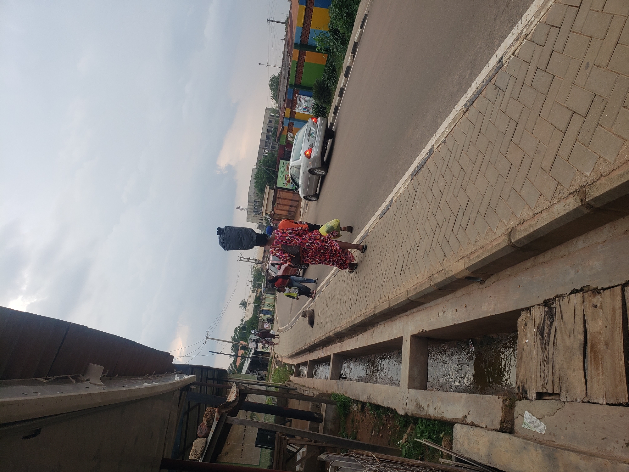 The image depicts a street scene in Narayi, Nigeria. A woman is walking on a paved sidewalk, carrying a large black bag balanced on her head. She is wearing a colorful, patterned dress and a backpack. To her right, a silver sedan is driving on the road. Several other people are visible walking in the distance on the sidewalk, and a dog is lying down near them. The sidewalk is bordered by a concrete drainage ditch filled with water on the left side of the image. In the background, there are buildings, fences with colorful painted sections, and utility poles with wires. The sky is overcast with clouds. The overall environment suggests a moderately populated urban area with typical infrastructure for a Nigerian city.