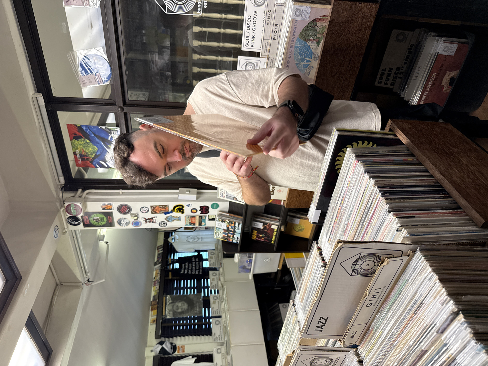 A medium shot captures a man in a record store, inspecting a vinyl record. He stands amidst rows of organized records, categorized and labeled as "JAZZ" and "SOUL/DISCO FUNK/GROOVE". The store, identified by "bungkumhouse records" labels, features shelves packed with records and merchandise. A window displays stickers and posters, while clothing is visible in the background. The location is inferred to be Bangkok, Thailand, based on the "bungkumhouse" label.