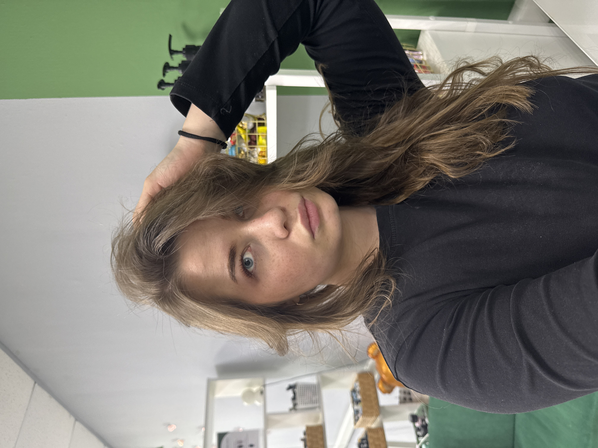 A woman is positioned in front of the camera, with her head angled slightly to the left. She has light brown hair and blue eyes. She is wearing a black long-sleeved shirt and has a black hair tie on her wrist. Her hand is resting on her head.
The background reveals elements of a room with white shelves containing various items, some identifiable as cosmetic or hygiene products. A green wall is visible at the top, with some black fixtures mounted on it.
The location appears to be indoors, potentially a room within a residence or a commercial space. There is no indication in the image that the location is in the city of Chaikovsky, Russia.