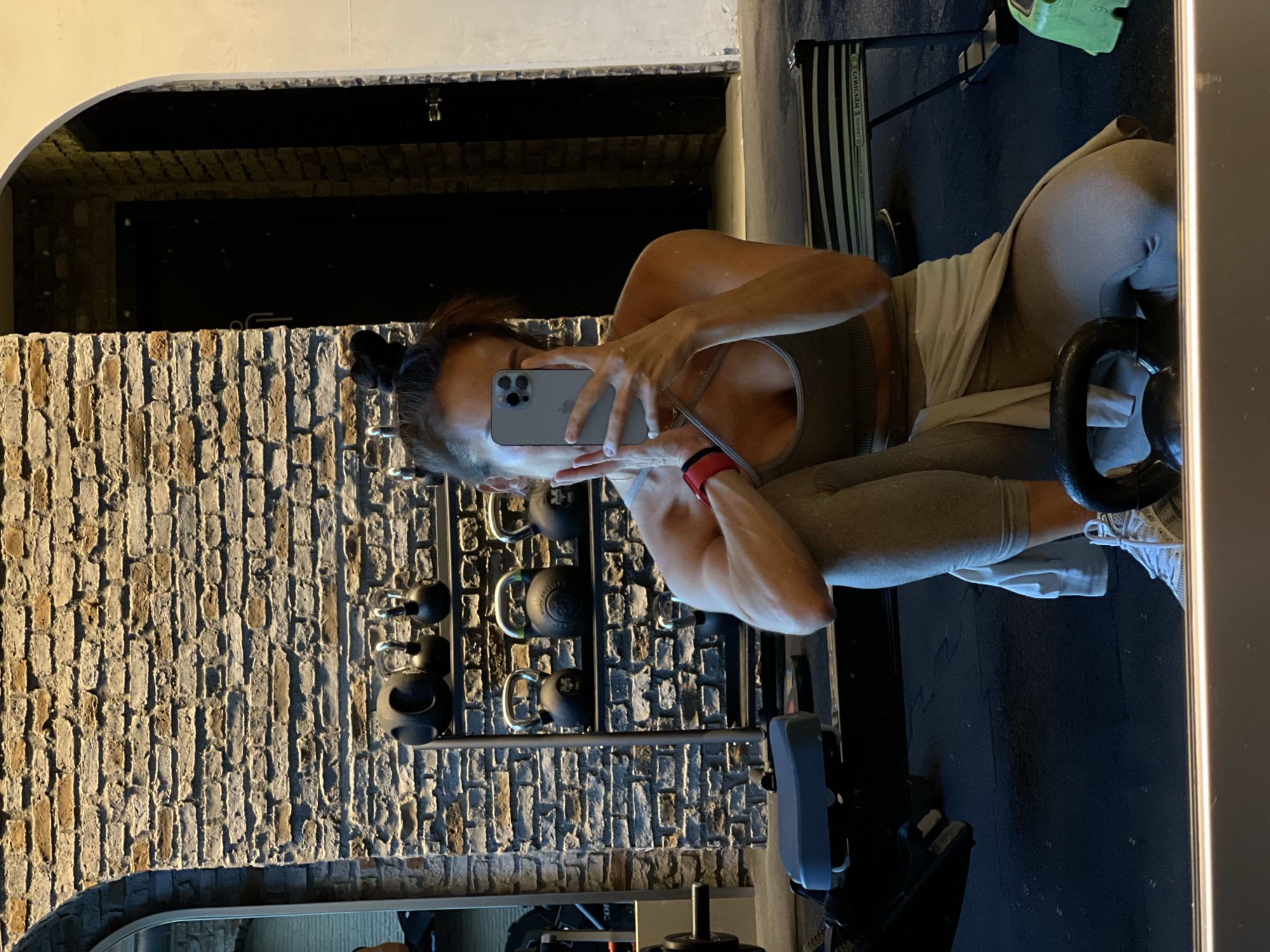 A person is taking a mirror selfie in a gym in Şişli, Turkey. The person is wearing athletic clothing and is holding a smartphone to take the photo. The gym environment includes brick walls, weight racks with kettlebells, gym equipment, and a mirror. The lighting in the gym appears to be artificial. The person's face is obscured by the phone.