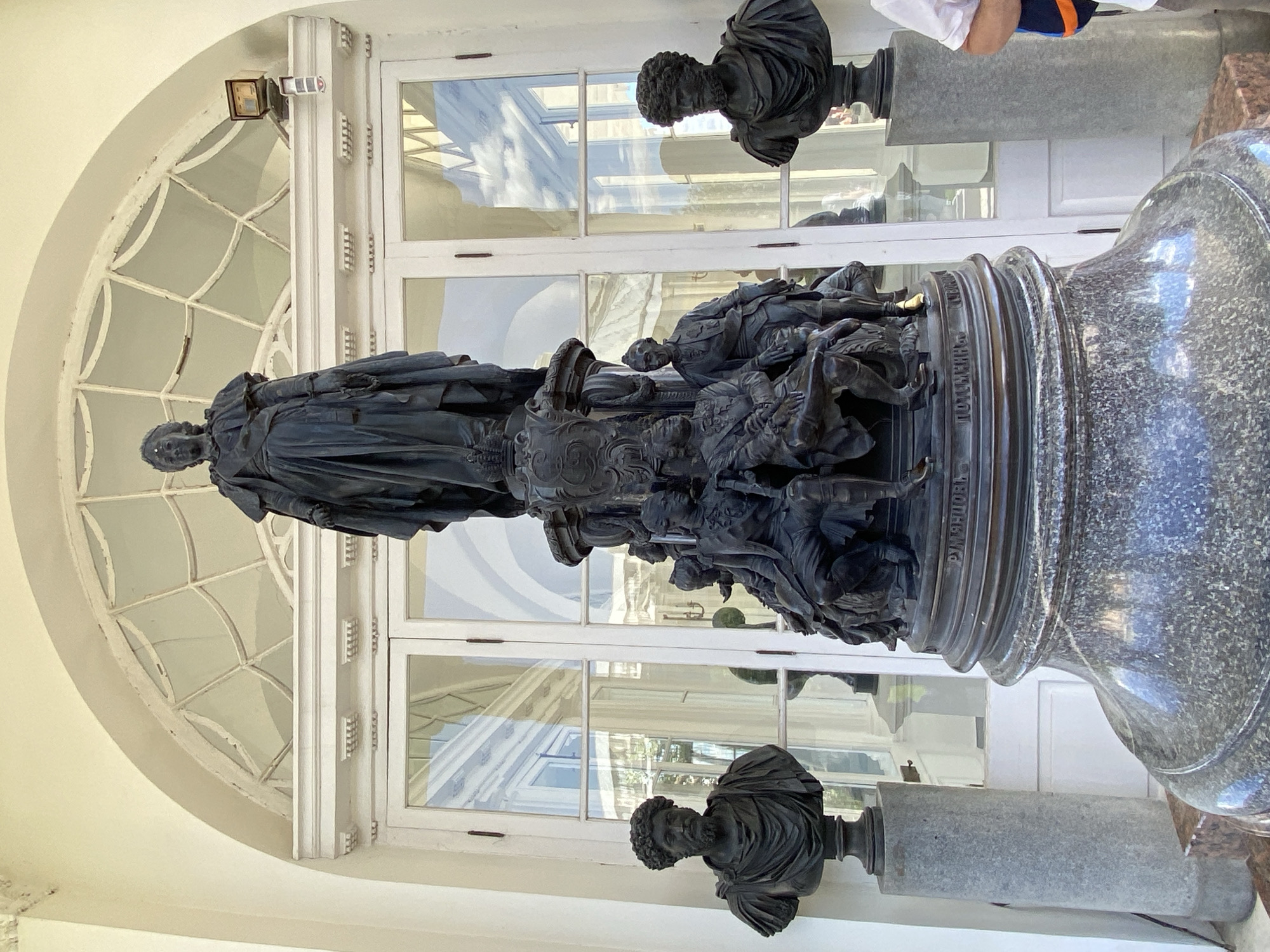 This image shows a bronze monument to Catherine the Great located in front of a large white arched doorway with glass panes in the city of Pushkin, Russia. The monument features a central statue of Catherine the Great atop a tiered base adorned with smaller figures and inscriptions. Flanking the monument are two black bronze busts on cylindrical pedestals. The bust on the left depicts a man with curly hair and a toga, while the bust on the right is similar, also showing a man with curly hair and draped clothing. In the background, the doorway leads into a brightly lit interior, and a person wearing a white shirt and a blue and orange striped bag is partially visible on the right side of the frame. The environment appears to be an outdoor area, possibly a palace or park setting, with reflections of clouds and architecture visible in the glass panes.