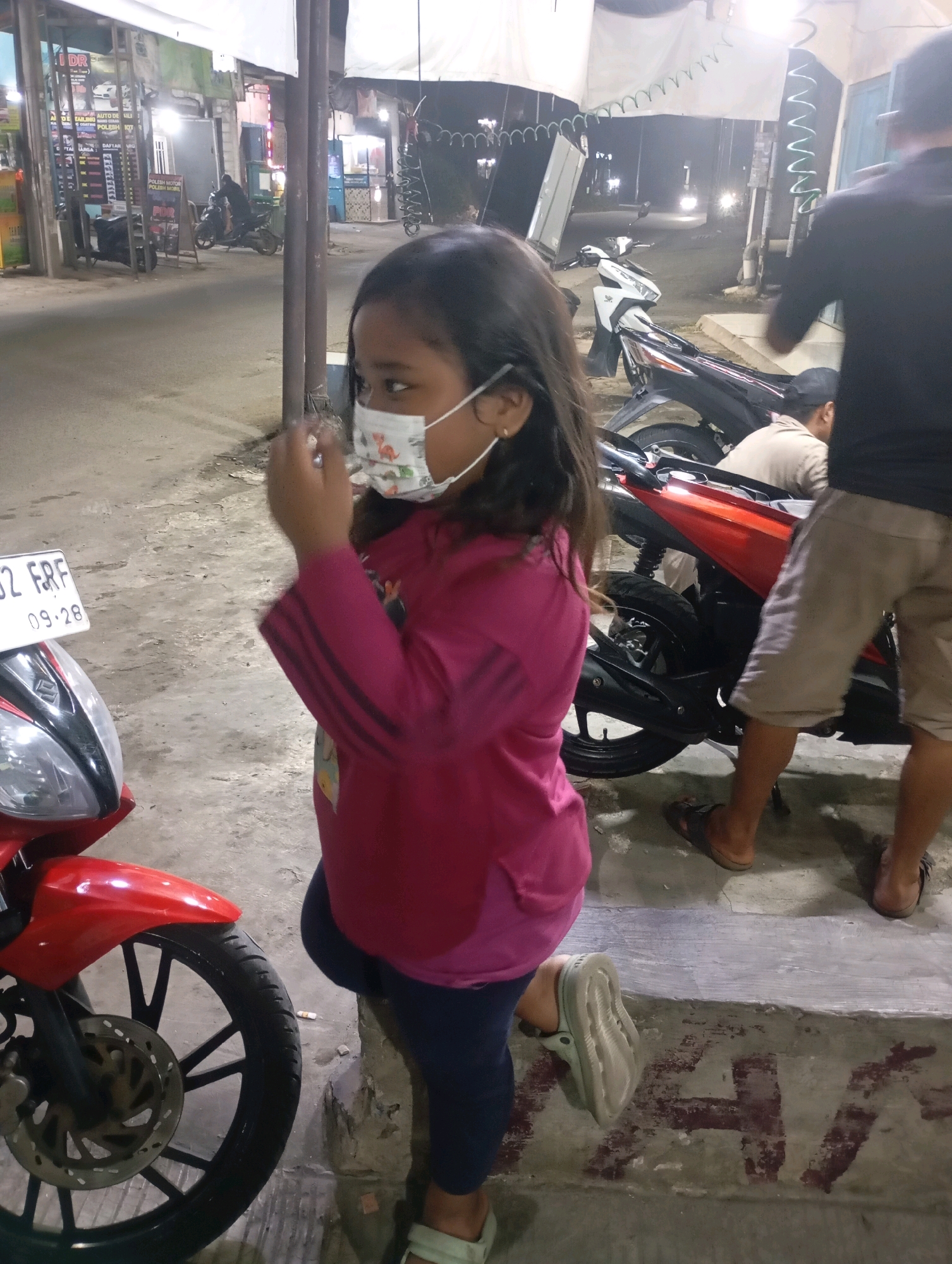 In Babelan, Indonesia, a young girl wearing a face mask with a dinosaur print is standing on a step. She has dark hair and is wearing a pink long-sleeved shirt and dark pants. Her right hand is near her face, touching the mask. To her left, a red motorcycle is parked, its front wheel and fender visible. Behind her, other people and motorcycles are present in an outdoor setting, possibly a street or market area with shops in the background. The ground is paved with concrete.