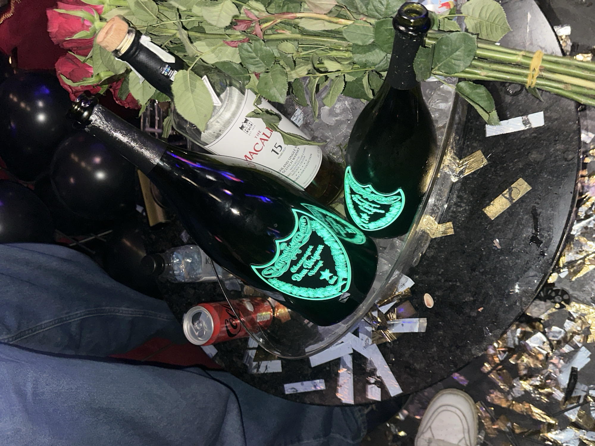 Here is a detailed description of the image:
**Scene:** An indoor scene, likely a celebratory setting. The focus is a glass-topped table cluttered with luxurious items and remnants of celebration. The location is identified as Unknown, Unknown.
**Objects:**
* Two bottles of Dom Perignon champagne with glowing green labels.
* A bottle of Macallan 15 Year Old scotch whisky.
* A can of Coca-Cola.
* A bouquet of red roses with visible green stems.
* Several black balloons.
* Confetti scattered around the table.
**People:**
* A person is partially visible, wearing blue jeans and white sneakers. Their legs are positioned next to the table.
**Environment:**
* The table appears to have a dark, speckled surface, likely granite or marble.
* The floor has a mix of light and dark areas with reflective confetti scattered around.
**Actions:**
* The scene suggests a recent celebration with alcohol, flowers, and confetti.
* The presence of unopened bottles implies the celebration may be ongoing or was recently concluded.