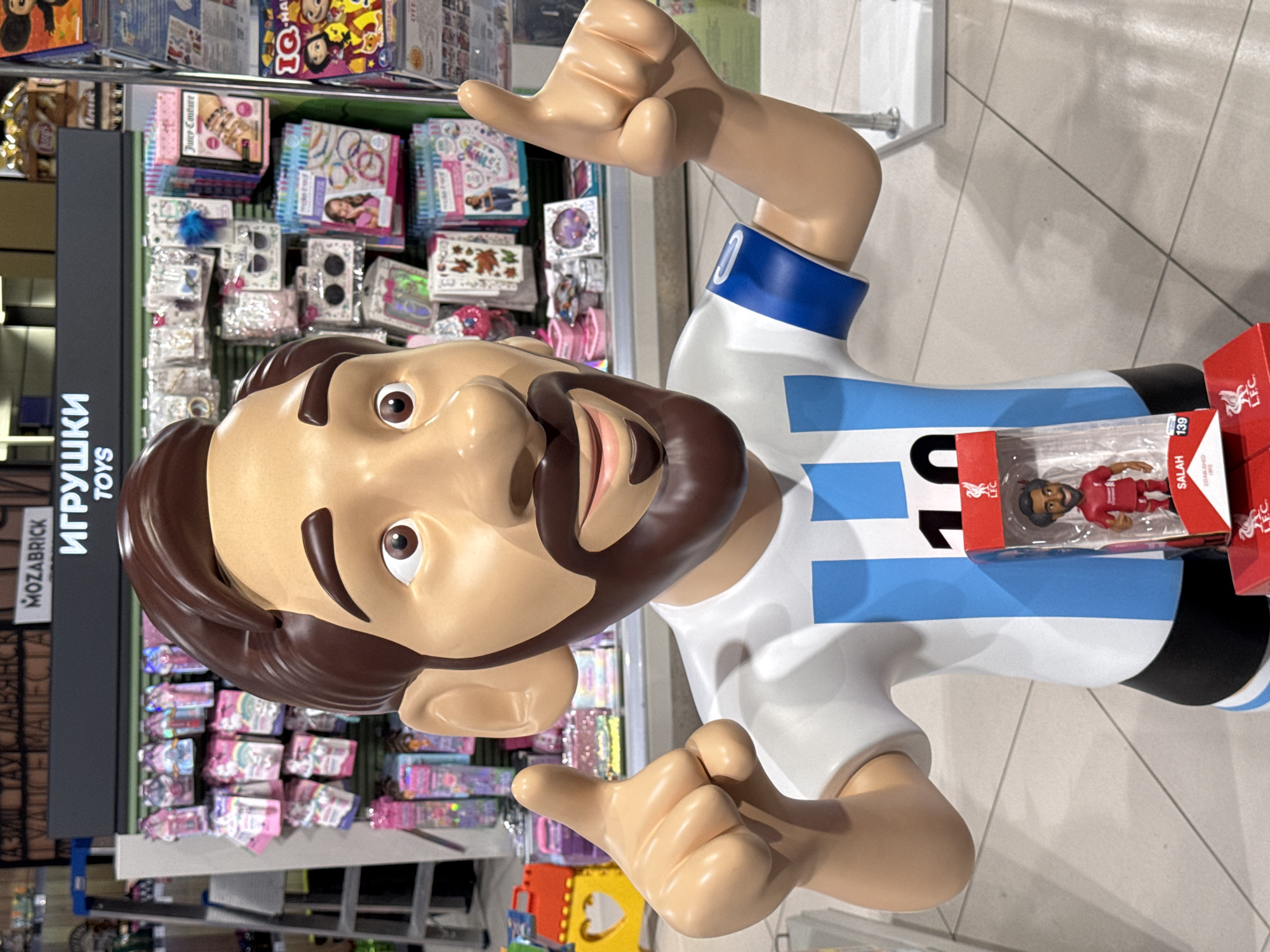 A cartoon figure with a beard and a white and blue striped shirt is in a store, in front of a shelving unit displaying various toys. The figure is giving a thumbs up with both hands. A toy figurine of "SALAH" in a red box sits on the figure's side. The shelving unit has a sign in Russian saying "Toys". The environment appears to be a well-lit retail setting, likely a toy store. The setting is likely in the city of Sochi, Russia, as "Тайм Маллавянск" is partially visible on the left side of the image.
