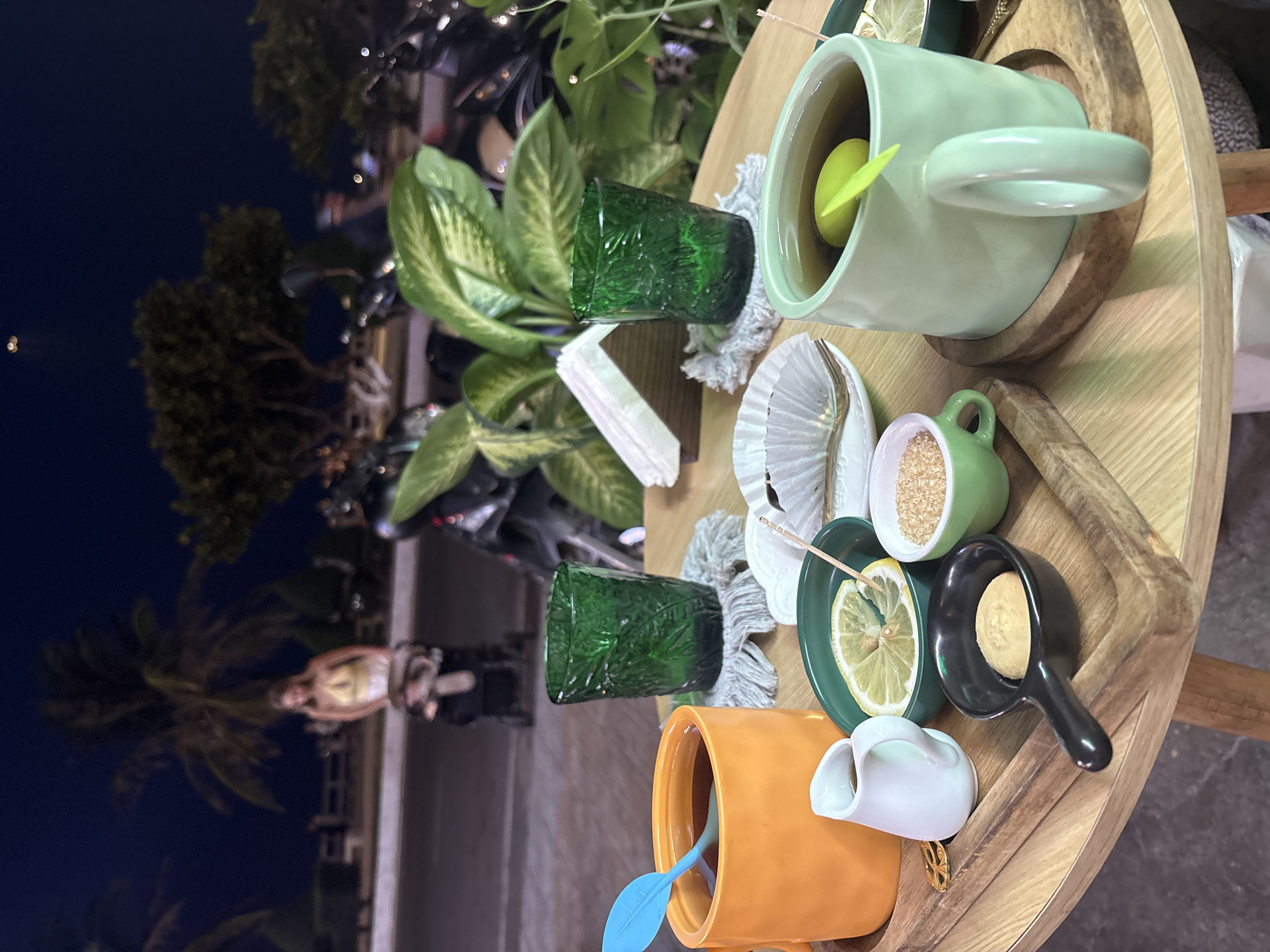 On a wooden table outdoors, presumably in Bắc Nha Trang, Vietnam, given the presence of tropical flora and a dimly lit cityscape in the background, an assortment of tea-related items is arranged. There's a light green mug with a yellow lemon inside, a small green cup filled with brown sugar, a black spoon-shaped dish holding a cookie, and an orange mug with a blue stirring utensil. Adjacent is a small white creamer pitcher. Two textured green glasses, a seashell, and white napkins are also present. The light suggests a nighttime setting. The overall scene suggests a tea service or setup.