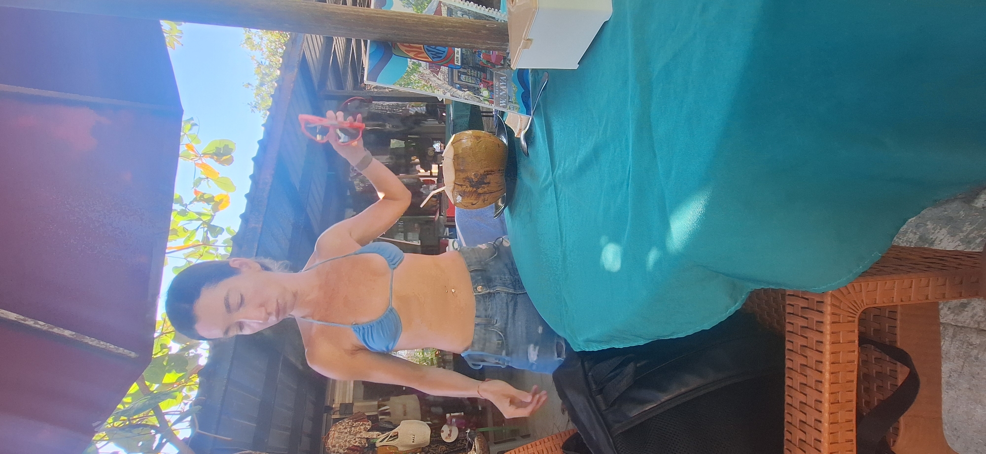 A woman stands at a table covered with a teal tablecloth. She is wearing a blue bikini top and ripped denim shorts. In her right hand, she holds a pair of red sunglasses. On the table, a whole coconut with a straw is placed on a black plate. A menu with text that reads "WARUNG W. NAYA" is visible to the right of the coconut. The scene appears to be outdoors, under a thatched roof structure, with sunlight filtering through trees and illuminating the area. Various bags and items are displayed on shelves in the background, suggesting a market or shop setting. A black backpack rests on a wicker chair to the left of the table.