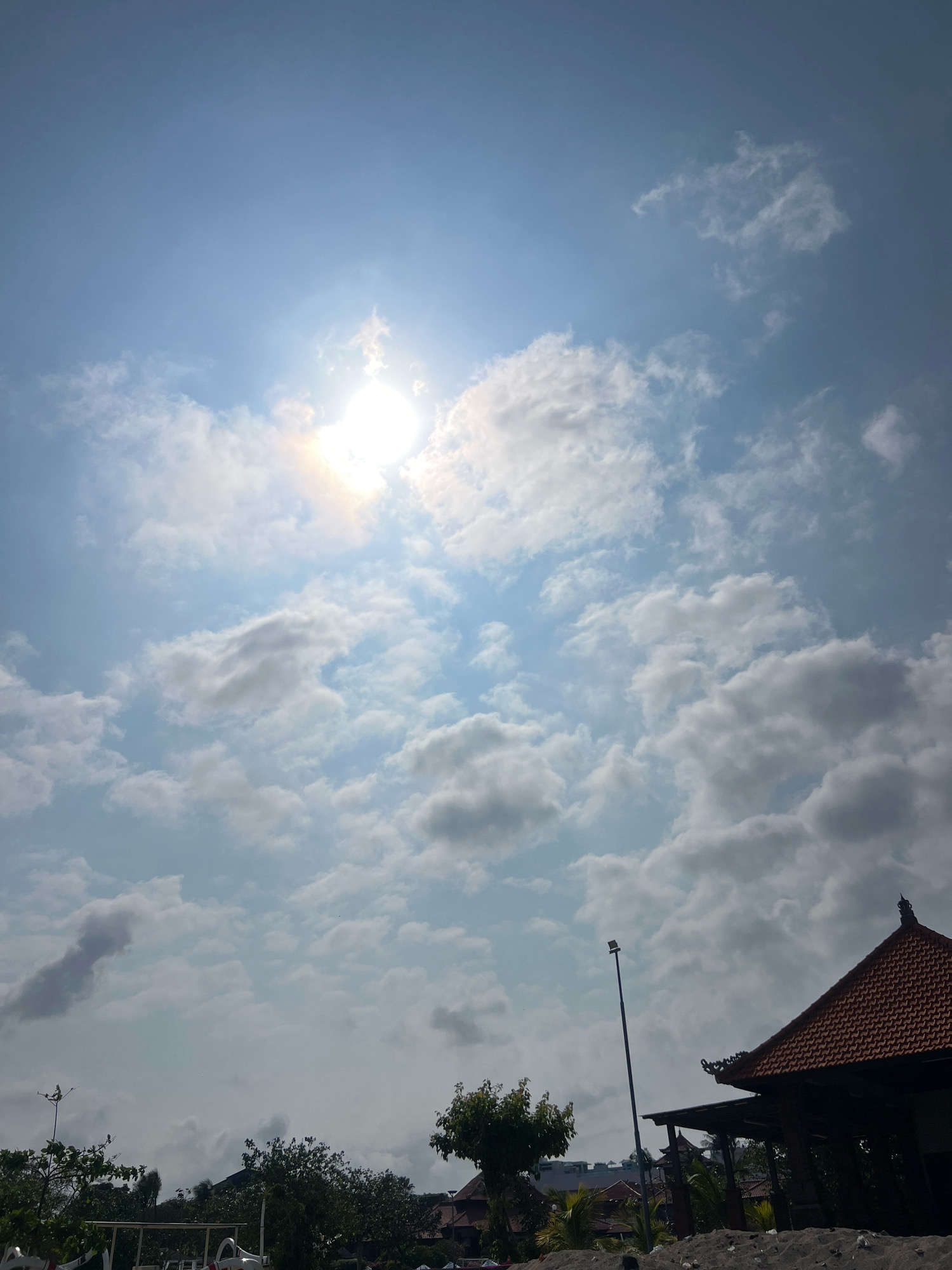 The image depicts a bright, sunny day with scattered clouds in the sky, a scene typical of Kuta, Indonesia. The sun is high and appears to be the primary light source, casting a bright glow. In the foreground, the image is dominated by clouds, some appearing fluffy and white, others darker and more diffused. To the right, a traditional Balinese-style building with a red tiled roof is partially visible, suggesting a resort or temple structure. A tall, slender lamppost stands near the building. Below the sky and building, trees and some white structures are visible, possibly part of a beachside establishment or garden. The overall impression is one of a clear day with a touch of tropical architecture. There are no people visible in the frame.