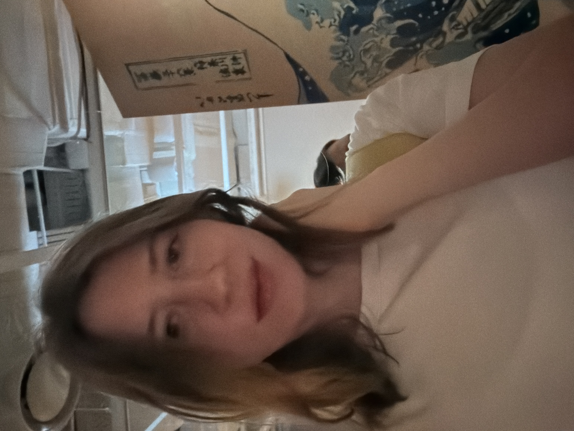 The image shows a young woman with light brown hair, wearing a white t-shirt, holding her hand to her neck. She is taking a selfie in an indoor environment. Behind her, a large artwork with Japanese characters and a wave motif is visible on a light-colored surface. The ceiling features exposed pipes and lighting fixtures, suggesting a commercial or public space. A second person's head and shoulder are partially visible in the background to the right. The lighting is soft, and the image appears to be taken with a phone camera. The location is not specified within the image, and there is no visual information to determine it is in Seoul, South Korea.