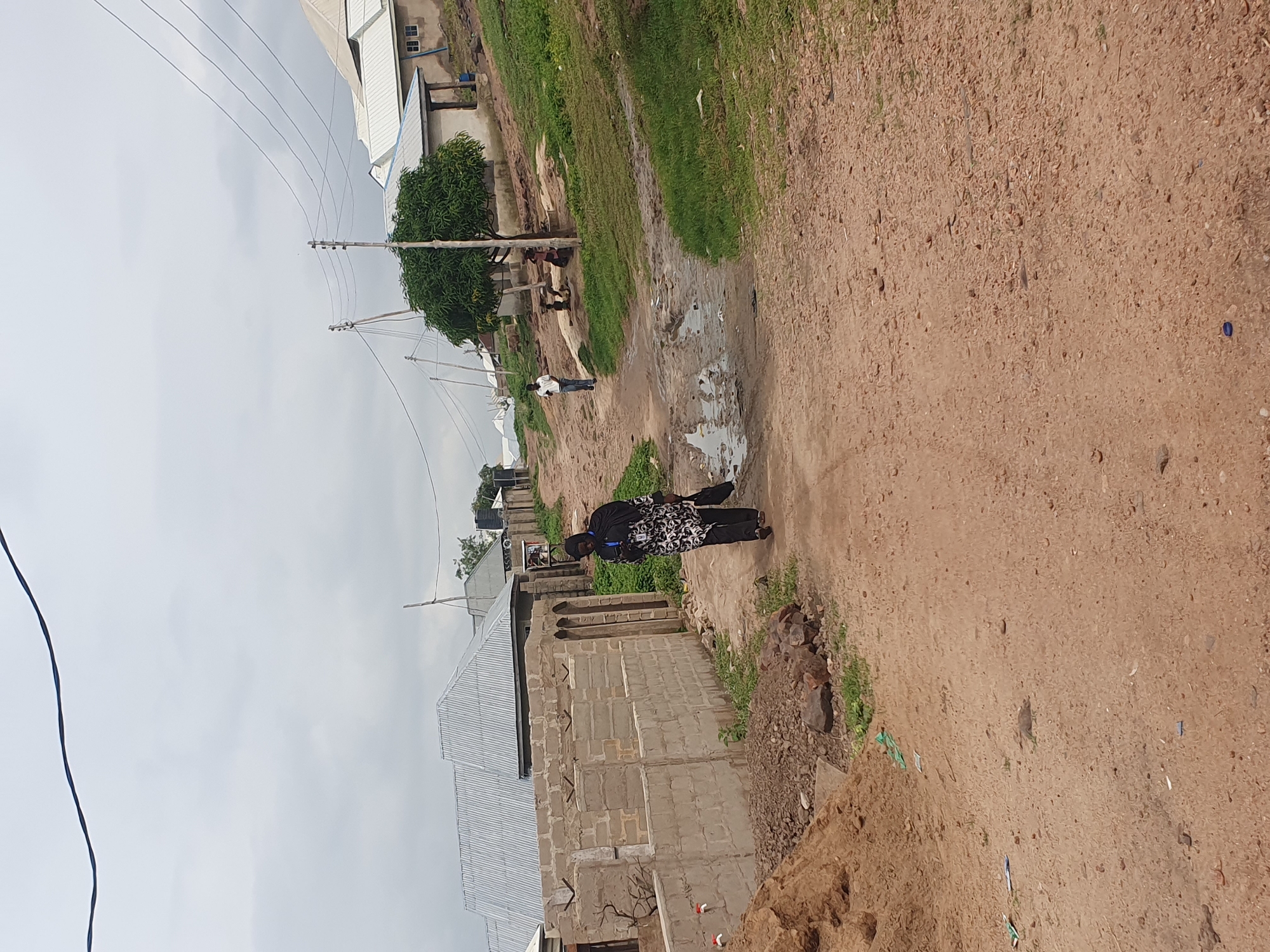 The image shows a dirt road with puddles in a residential area in Lokoja, Nigeria. A woman wearing a black hijab and a patterned dress walks away from the camera, carrying a black bag. She is on the right side of the road, near a partially constructed brick wall. In the background, several houses with corrugated metal roofs are visible, along with trees and utility poles. Two men and some children are seen further down the road, some sitting under a tree. The sky is overcast. The ground is uneven, with patches of grass and exposed soil. Debris and a blue bottle cap are scattered on the road.