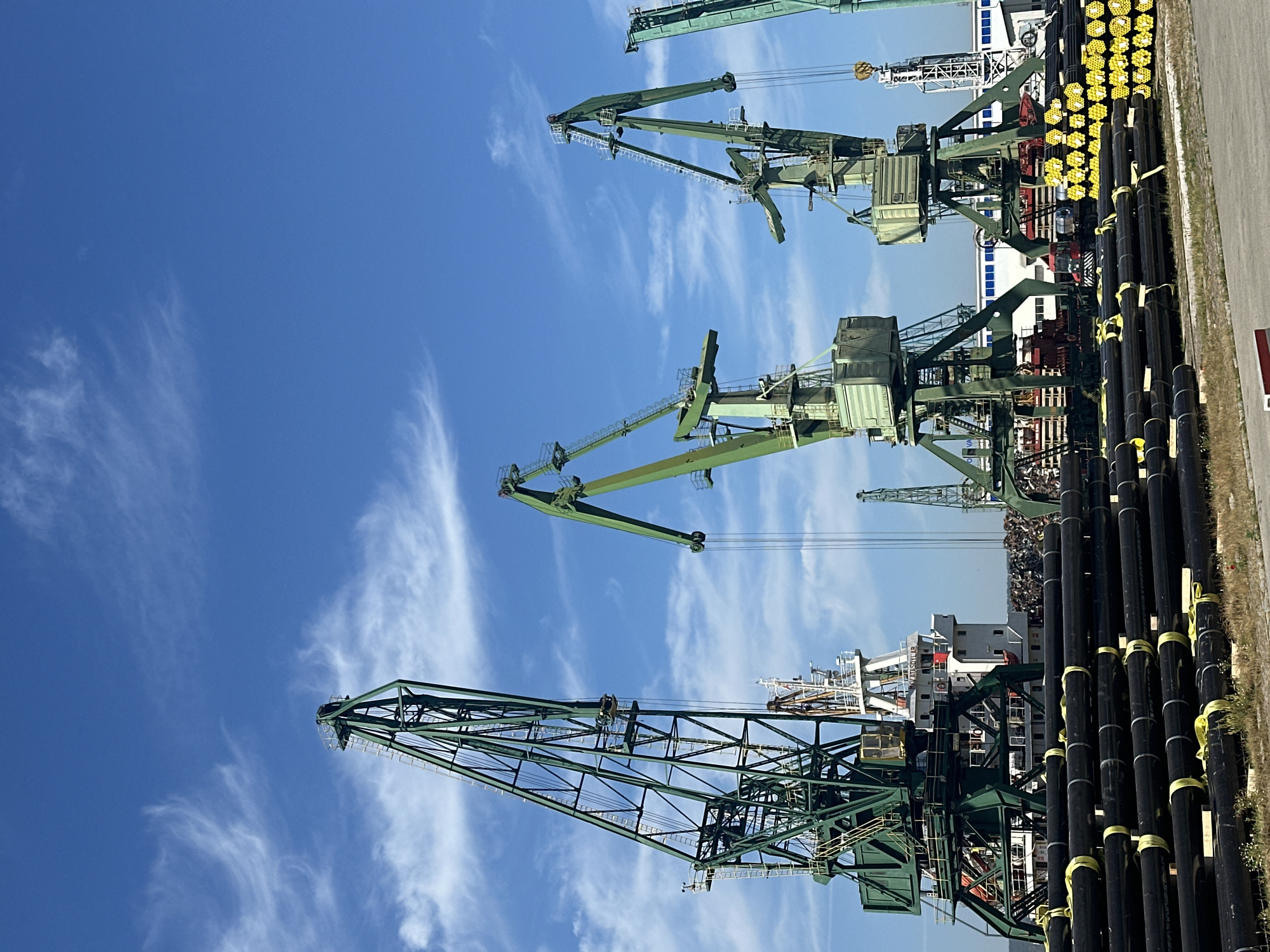 In an industrial port setting under a bright blue sky with wispy clouds, a large stack of black pipes with yellow straps fills the foreground. Behind the pipes, a row of green harbor cranes dominates the mid-ground. One crane on the far left is particularly tall and extends high into the sky. Further back, a large white ship with red accents is docked, and beyond it, a large pile of scrap metal is visible. Another cluster of yellow-capped pipes is stacked to the right of the green cranes. The scene suggests active maritime and industrial operations.