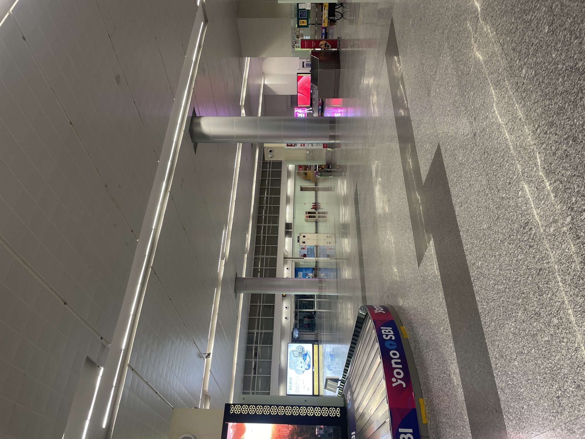 The image depicts the interior of a large, modern airport terminal, likely Raipur Khurd, India, given the presence of "Yono SBI" branding on the baggage claim carousel. The space is characterized by its expansive, polished granite floor, reflecting the bright, diffused lighting from long, linear fixtures embedded in the suspended ceiling. Several large, cylindrical grey pillars support the ceiling and divide the open space.
In the foreground, a baggage claim carousel is prominently visible on the left, featuring bold "Yono SBI" branding in pink and blue. To the left of the carousel, a large digital display screen shows a vibrant image of a cityscape at sunset.
The midground shows a wide, empty expanse of the terminal floor, leading towards a series of entrances and information displays. A large digital billboard on the left displays an advertisement for "RAN BAAS" with imagery of hotel rooms and amenities. Further back, the terminal appears to have a section with large windows and what looks like reception desks or service counters. There are a few people visible in the distance, appearing as small figures near the entrances, suggesting low passenger traffic at the time of the photograph.
The environment is clean and well-maintained, typical of an airport terminal. The lighting is bright and even, creating a sense of openness and spaciousness. The overall impression is one of a modern, functional transportation hub.