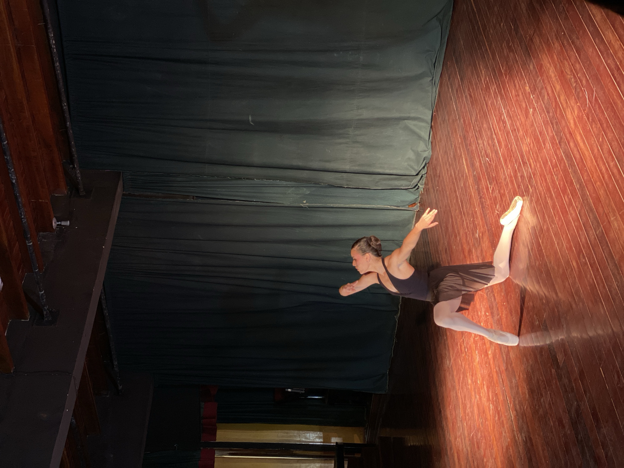The image shows a ballerina mid-movement on a stage, likely during a performance or rehearsal. She is wearing a dark leotard and tights with ballet slippers. The backdrop is a dark, draped curtain, and the stage is made of wooden planks. The overall lighting suggests a theatrical setting. There is no information in the image to confirm the location is in Juan Griego, Venezuela.