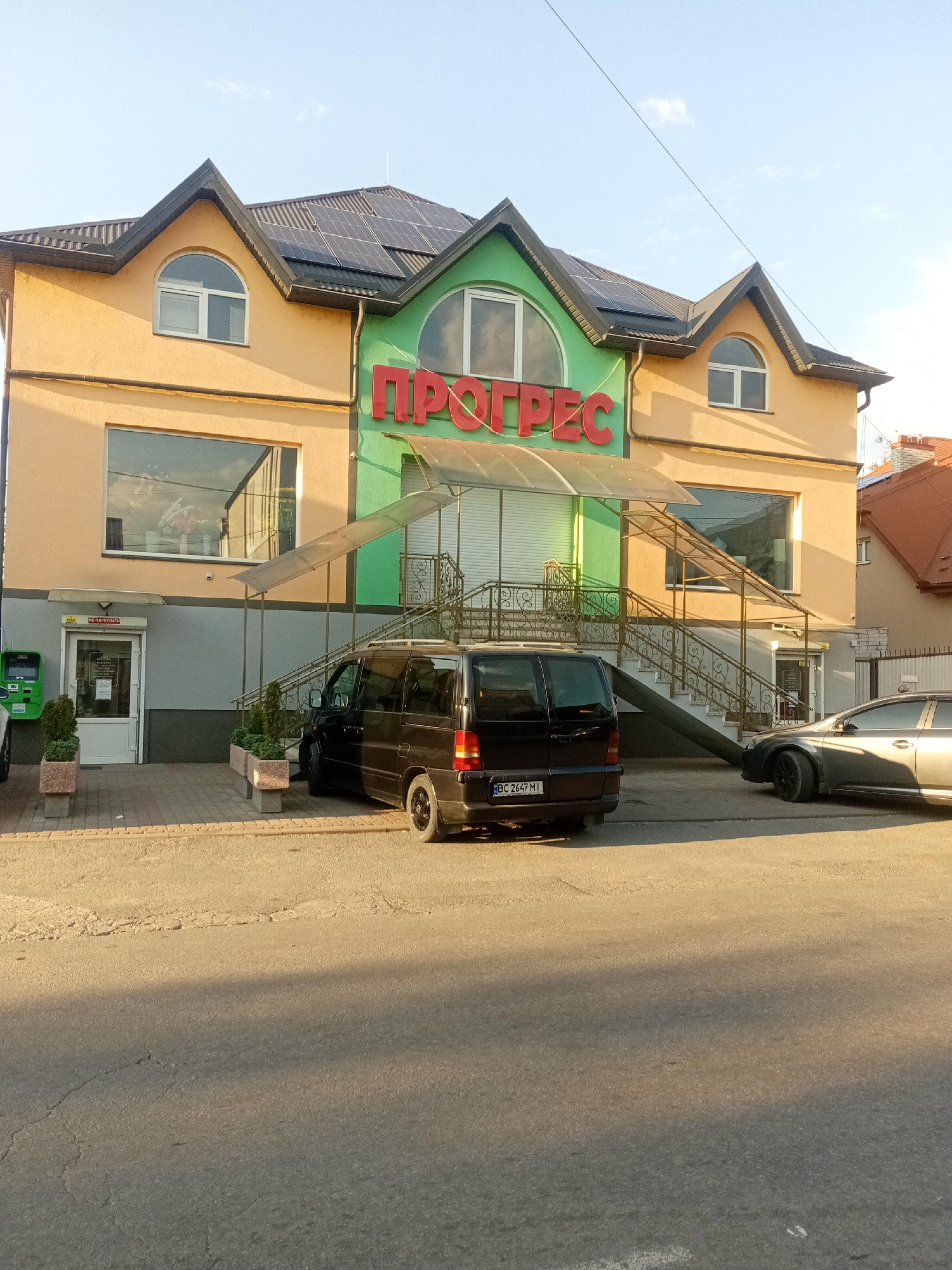 The image shows the facade of a building with the word "ПРОГРЕС" (Progress) in red letters above the entrance. The building has an orange and green color scheme and appears to have solar panels on its roof. In front of the building, there is a black van with the license plate "BC 2647 MI" parked on the right side. To the right of the van, a silver car is parked. A covered walkway with stairs leads up to the entrance. The scene is set on a street with asphalt pavement and includes some potted plants and a payment terminal. The overall setting suggests a commercial establishment in a city.