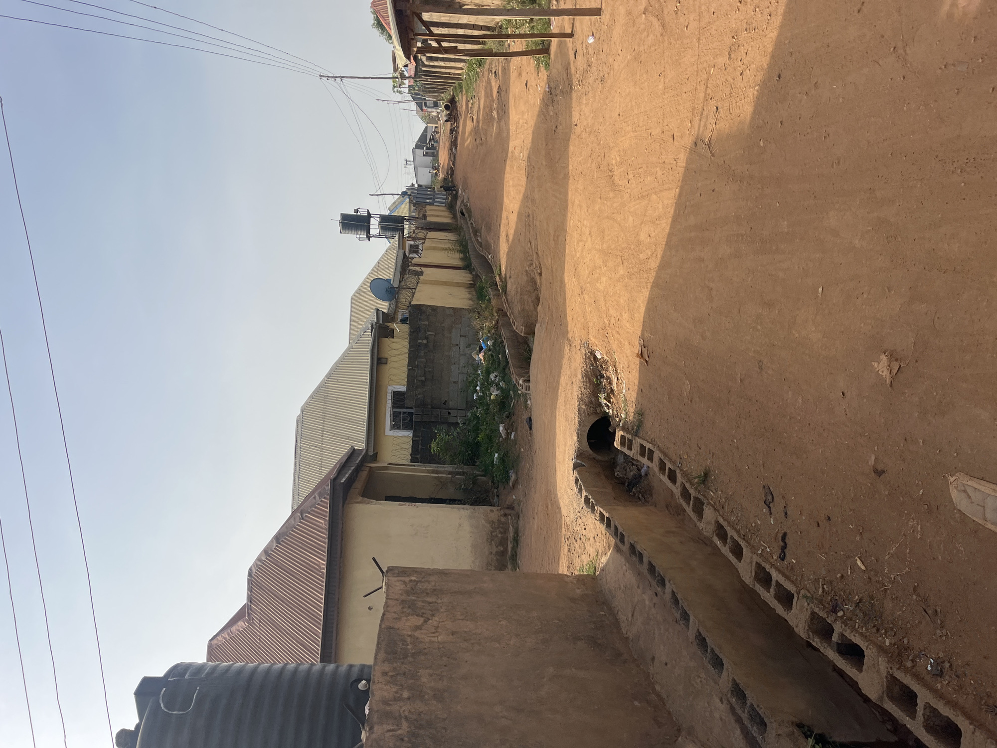 The image captures a static, unpaved, reddish-brown dirt road or pathway in what appears to be a semi-urban or residential area in Abuja, Nigeria. The photograph is taken during daytime under a clear, bright sky, indicated by the distinct shadows cast across the path from the right.
Along the right side of the path, a concrete drainage ditch, constructed from hollow concrete blocks, runs parallel to the dirt ground. The ditch contains some debris and sparse vegetation. The ground of the path itself shows minor undulations, tire marks, and scattered small stones or aggregates.
To the left of the path, several buildings are visible. Most feature corrugated metal roofs, common in the region. One prominent building is painted yellow, with a small window and what appears to be a partially exposed brick wall section. A large black water tank is seen in the foreground on the far left, while another smaller, gray water tank is mounted on a metal stand atop a building further down the road. A satellite dish is also affixed to one of the buildings. Overhead utility wires crisscross the bright sky, connecting to unseen poles.
No people or vehicles are present, and no specific activity or event is taking place. The scene portrays a quiet moment in a local street. There is no visible text.