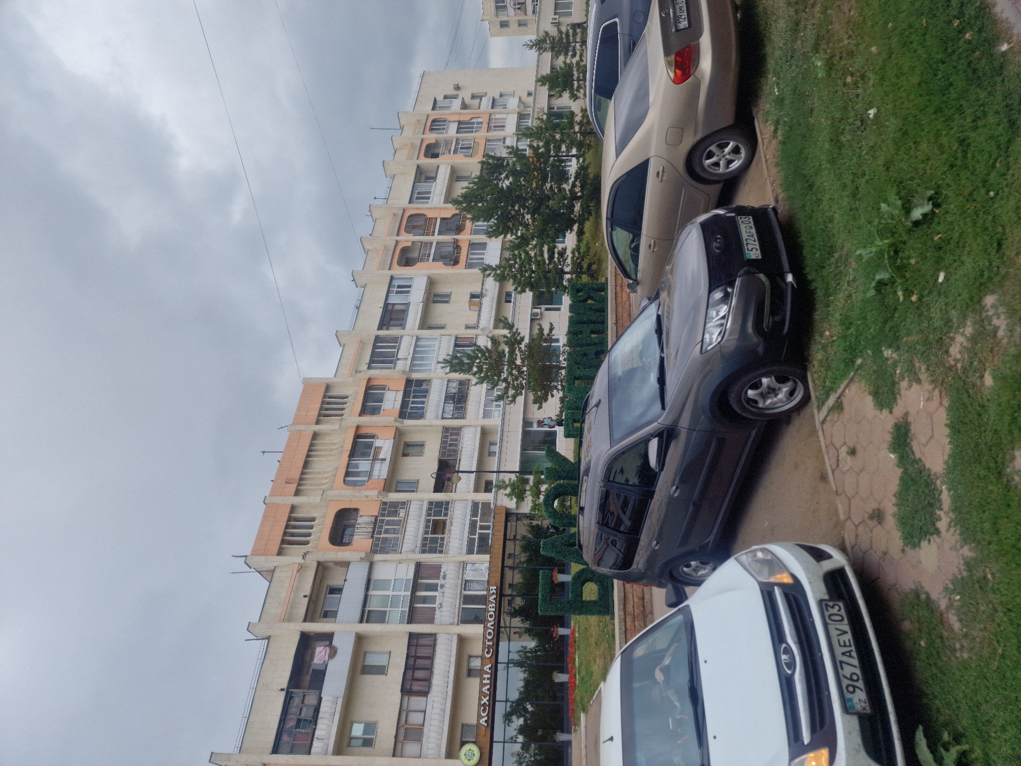 In Kokshetau, Kazakhstan, a cloudy day is captured with a street view. In the foreground, three cars are parked: a white sedan on the left with license plate KZ 967AEV 03, a dark gray minivan in the center with license plate KZ 572AFG 03, and a gold sedan on the right with license plate KZ 129AHK 03. Behind the cars, a large, multi-story apartment building dominates the scene. The building's facade features a mix of cream and peach-colored sections with visible balconies and numerous windows. In front of the building, a green, sculpted hedge forms large letters spelling out "БАКШЫ" in Cyrillic. Below the hedge letters, a sign with text in Cyrillic reads "АСХАНА СТОЛОВАЯ." Several evergreen trees are planted between the building and the parked cars. The ground is a mix of paved walkway and green grass. The overall atmosphere is overcast, suggesting potentially inclement weather.