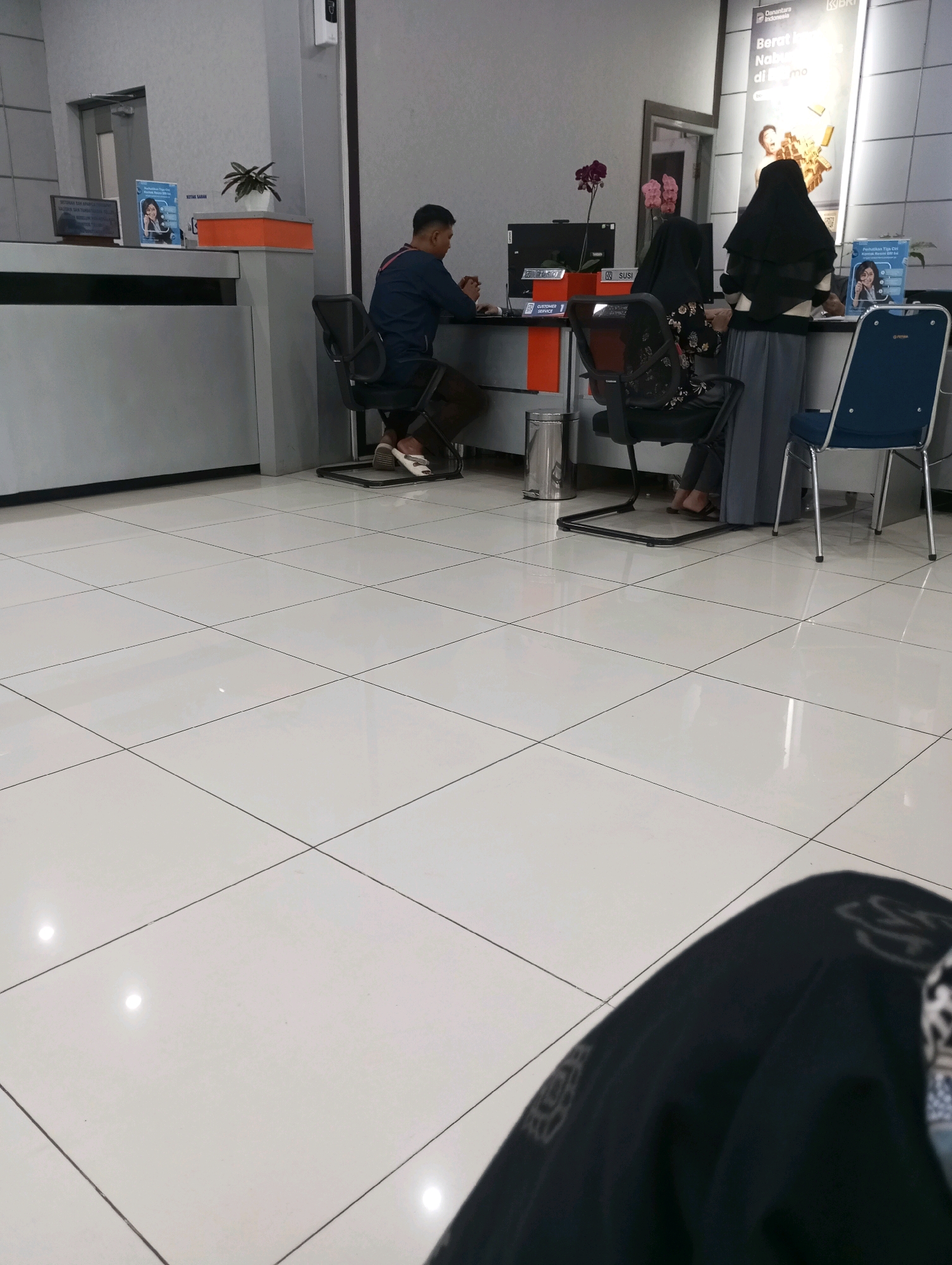 The image depicts the interior of a bank branch in Padang Panjang, Indonesia. The scene is set in a modern, well-lit space with a tiled floor. In the foreground, a person is seated, with only their legs and lower torso visible, suggesting they are a customer or visitor. In the middle ground, a male customer, wearing a dark blue shirt and brown trousers, is seated at a counter, facing a computer monitor. Behind the counter, two female employees are visible. One, wearing a black hijab and patterned top, is standing and interacting with a customer. The other, also in a hijab, is seated and appears to be working. On the counter, there are computers, brochures, a plant, and a trash bin. The overall impression is one of a typical banking transaction environment.