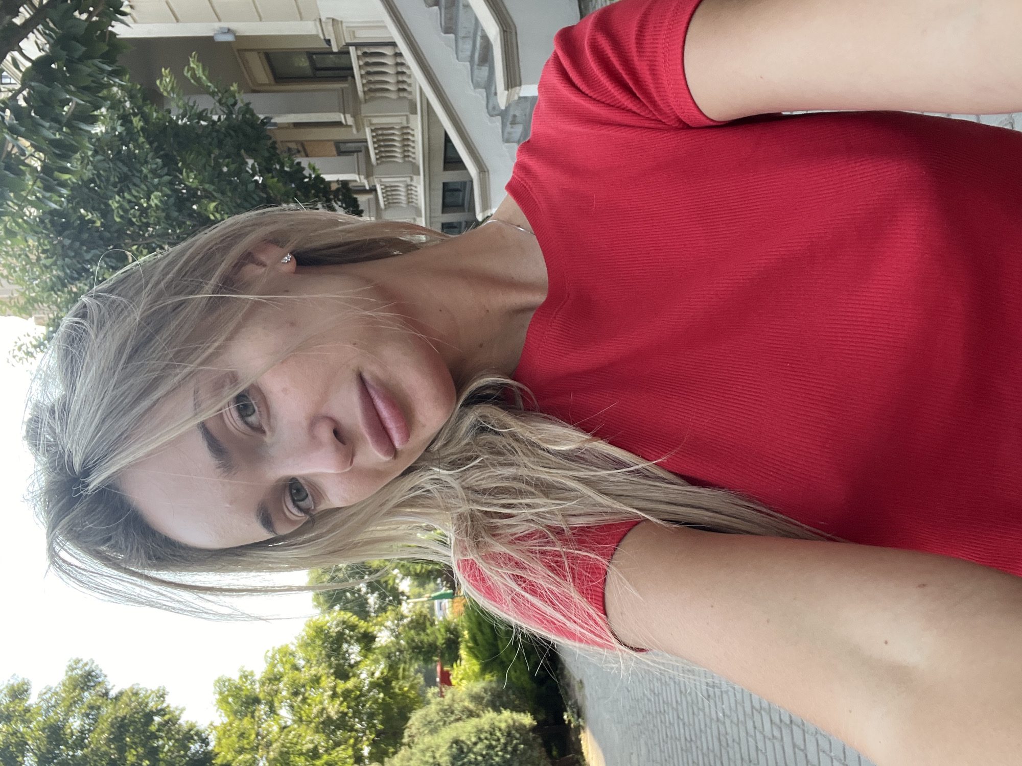 A young woman with long, blonde hair is taking a selfie outdoors. She is wearing a red, ribbed t-shirt and a thin silver necklace. Her hair is slightly messy, with some strands falling around her face. She is looking directly at the camera with a neutral expression. Behind her, there is a building with ornate architecture, including columns and a grand staircase. Lush green trees and bushes frame the scene. The ground in front of her appears to be a paved walkway. The image is lit by natural daylight.