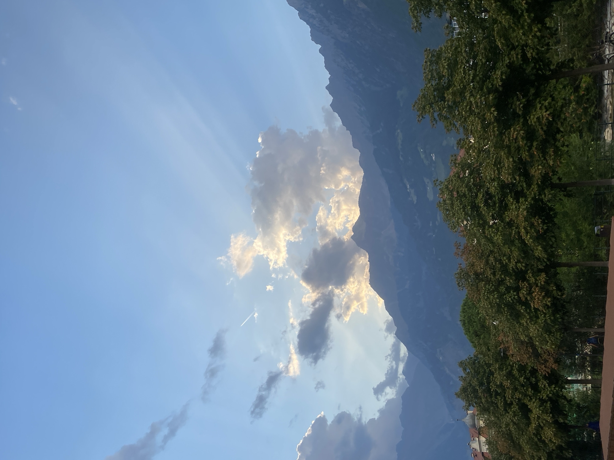 The image captures a view of the mountains surrounding Meran, Italy, under a partly cloudy sky with crepuscular rays visible. In the foreground, a line of mature trees with green foliage borders a pathway or park area. A red brick structure and a domed building are partially visible to the left behind the trees. A faint contrail from an aircraft streaks across the sky. The mountains are densely forested and layered, with the furthest ranges appearing in shades of blue and gray due to atmospheric perspective. The lighting suggests it is either sunrise or sunset, with warm light illuminating some of the clouds and mountain peaks.
