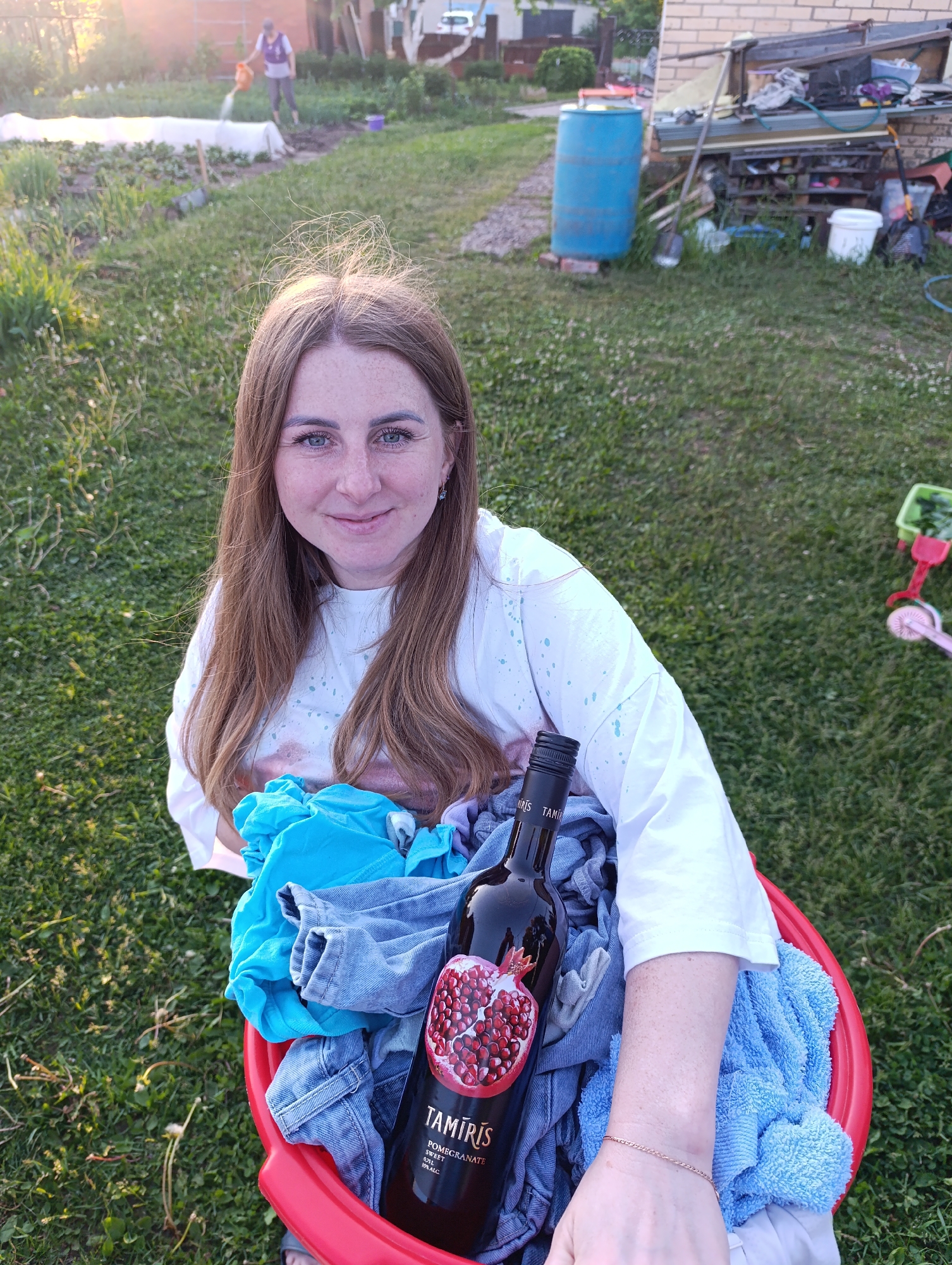 An outdoor scene depicts a light-skinned adult female positioned in the lower center foreground, illuminated by late afternoon or early evening golden sunlight. She has long light brown hair with subtle highlights, blue-green eyes, and freckles across her nose and cheeks. She wears a loose-fitting white t-shirt with light blue splatter patterns and a small silver-colored earring in her left ear. She smiles gently while looking directly at the viewer.
She holds a red plastic bucket containing folded clothing, specifically a light blue fabric item, denim jeans, and a light blue terry cloth towel. A dark-colored bottle of 'TAMIRIS POMEGRANATE SWEET WINE' (10% ALC, 0.7L) with a pomegranate illustration on its label rests among the clothing. Her right forearm, adorned with a delicate gold-colored chain bracelet, is visible on the right side of the bucket.
The scene is set in what appears to be a residential garden or backyard area in the city of Unknown, Unknown. In the midground, green grass with interspersed white flowers covers the ground. Another adult individual, wearing a purple and white top and light-colored pants, is visible in the upper left background, engaged in watering a garden bed with an orange watering can. The garden bed contains several rows of plants, some covered with white protective fabric tunnels.
To the right of the garden bed, a blue plastic barrel rests on bricks near a multi-level shelving unit made of wood and metal, piled with miscellaneous items including lumber, tools (a shovel, rakes), and white plastic buckets, all set against a light-colored brick building. A pink and green child's toy, resembling a small lawnmower or wagon, is situated on the grass in the lower right background. Additional blurry vegetation and a faint white vehicle are visible in the far background.