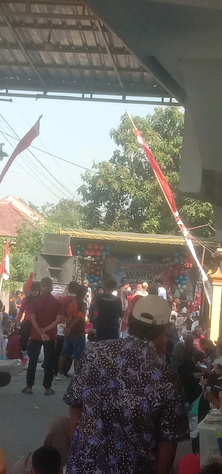 A group of people are gathered in an outdoor setting, possibly a community event, in Temuwulan, Indonesia. The scene is dominated by a stage or tent structure with a banner that reads "JALAN SEHAT SENAM SEHAT" and "DUSUN CANGKRI". Red and blue balloons are decorating the structure. Several individuals are standing or sitting in front of the stage, some appearing to be performers or organizers, while others are onlookers. Two Indonesian flags, red and white, are prominently displayed, one on the left and another on the right side of the image. Trees and buildings are visible in the background, suggesting an urban or semi-urban environment. The overall atmosphere seems festive.