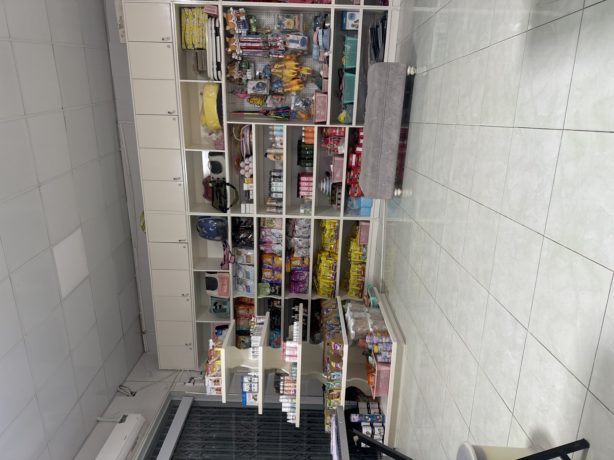 The image shows the interior of a pet supply store, likely located in Phu Thuy Ward, Vietnam. The store is well-lit with overhead fluorescent lighting. The walls are painted white, and the floor is tiled with large, pale gray square tiles.
The store features extensive shelving units filled with a variety of pet products. On the left, a multi-tiered display shelf holds numerous bottles, cans, and packages of pet food, treats, and grooming supplies. On the right, larger white shelving units are organized into compartments. These compartments contain an assortment of pet items, including bags of pet food, toys, leashes, collars, pet carriers, and grooming tools. Some shelves have pink and green baskets holding smaller items.
A grey upholstered bench with decorative legs sits on the tiled floor in front of the shelving units on the right. In the background, a window with a metal gate is visible, suggesting a storefront. An air conditioning unit is mounted high on the wall near the window. The overall impression is that of a clean, organized retail space dedicated to pet supplies. There are no people visible in the image.