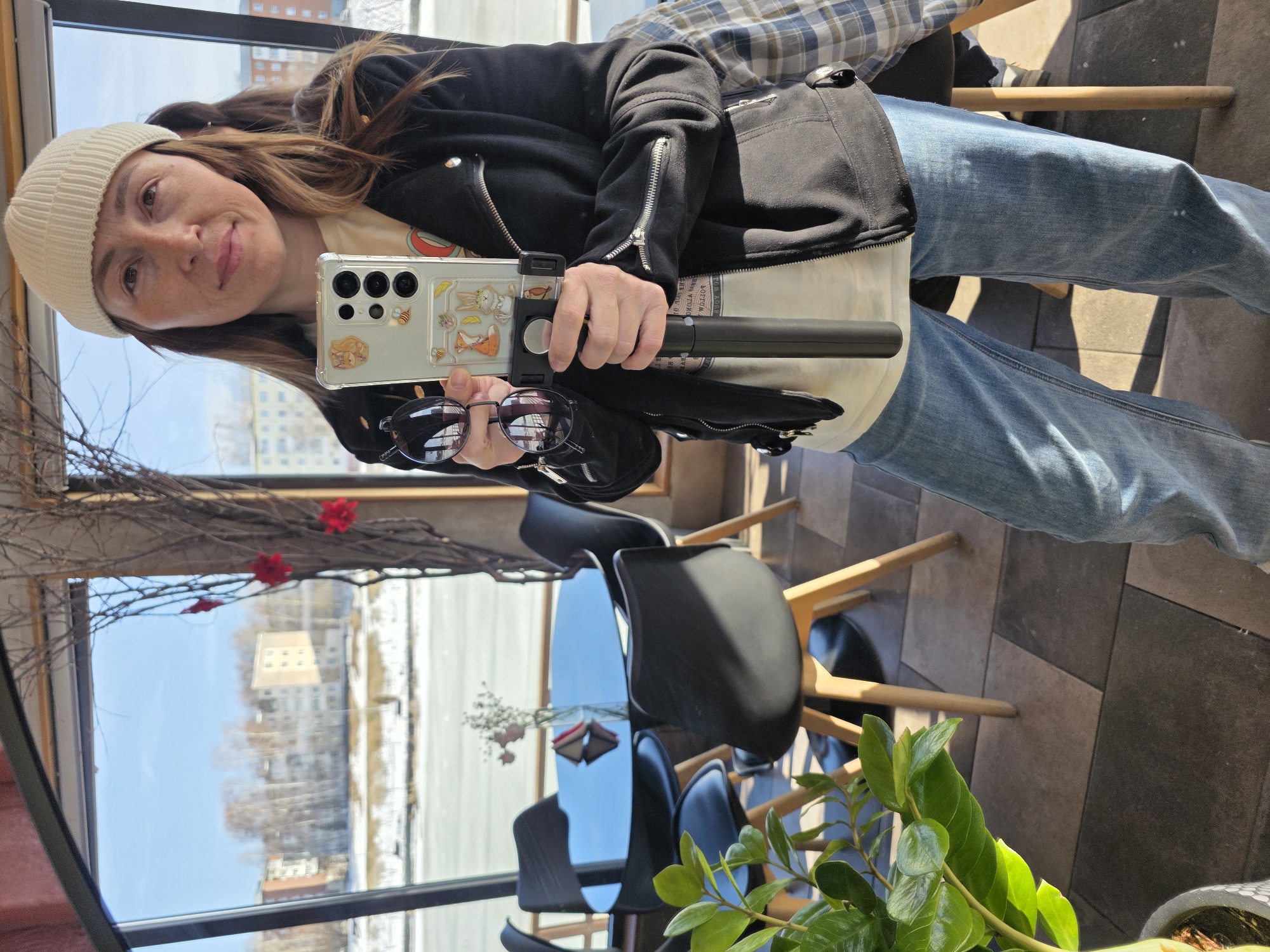 A woman is captured in a selfie, holding her smartphone attached to what appears to be a selfie stick or small tripod. The image is taken indoors, looking out through a large window, likely in a café or restaurant setting.
The woman, who appears to be in her 30s or 40s, has brown hair and is wearing a light cream or white knit beanie. She is smiling faintly and looking directly at the camera. Her complexion shows some freckles. She is dressed in a black zippered jacket, possibly a moto-style jacket due to the visible zippers on the sleeves and front. Underneath, a lighter-colored shirt (possibly cream or light yellow) with a graphic design is partially visible. She is also wearing blue denim jeans.
In her right hand, she holds a light-colored smartphone (possibly a Samsung Galaxy S24 Ultra given the camera module design) with a clear case adorned with cartoon animal stickers, including rabbits and foxes, and some bees. The phone is clamped onto a black selfie stick or tripod. Her left hand is resting near the phone, holding a pair of black-framed sunglasses, which reflect the window view behind her.
The indoor setting features a dark, reflective table and dark chairs with black seats and backs, supported by light-colored wooden legs. A small plant with vibrant green leaves is visible in the foreground on the right. Decorative branches with small red flower-like accents are draped across the top left portion of the window frame. The floor appears to be made of square or rectangular tiles in various shades of grey and brown.
Through the large window, a bright, clear blue sky is visible. The outdoor scene shows what appears to be a large body of water, possibly a river or lake, partially covered in ice or snow, stretching into the distance. Bare trees line the far bank, and several buildings, including a multi-story light-colored building, are visible in the background under the sunny sky. The overall impression is a bright, clear day, likely late winter or early spring given the bare trees and ice/snow.
